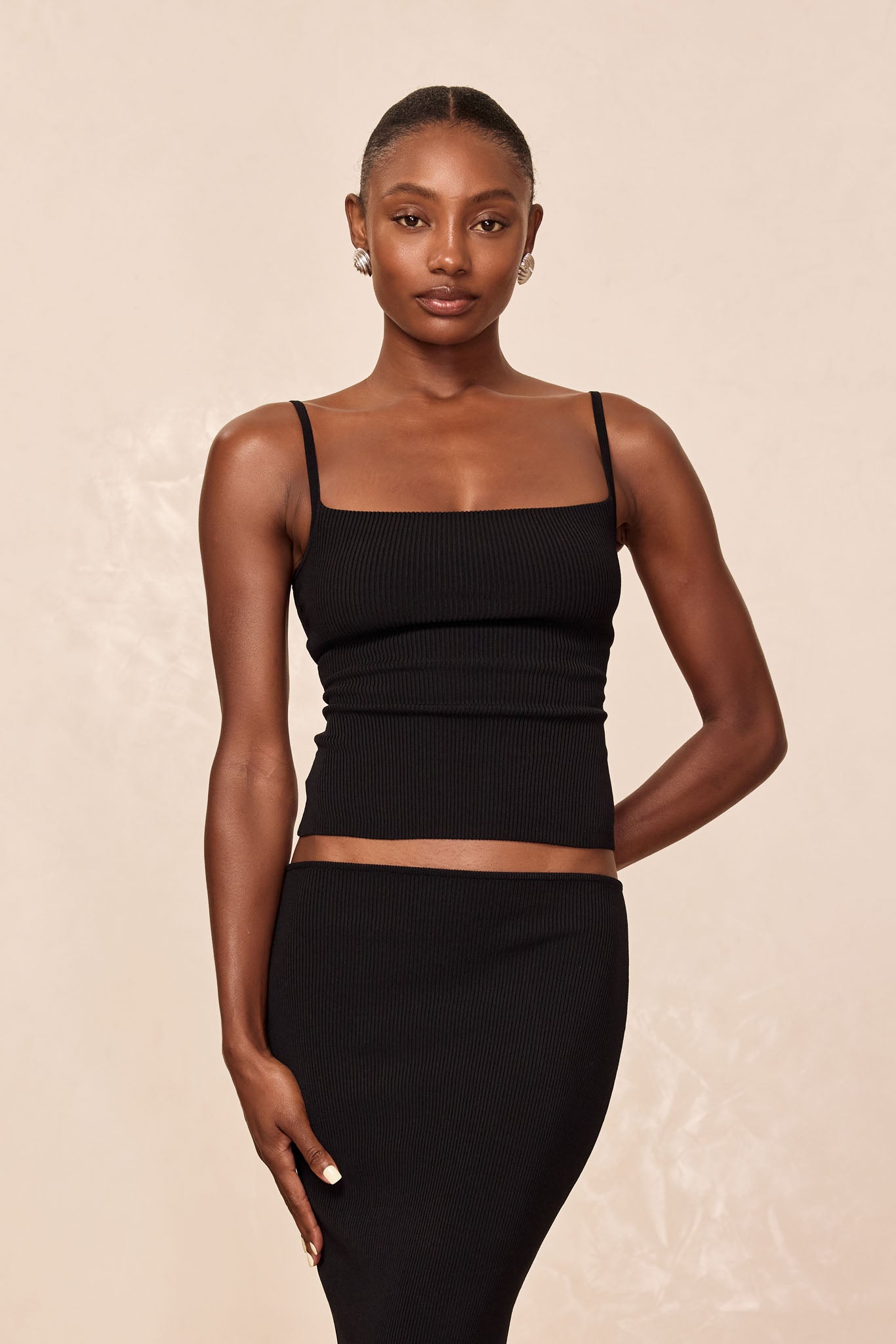 Model wearing black ribbed square-neck camisole and matching high-waist skirt against neutral background, hands at sides.