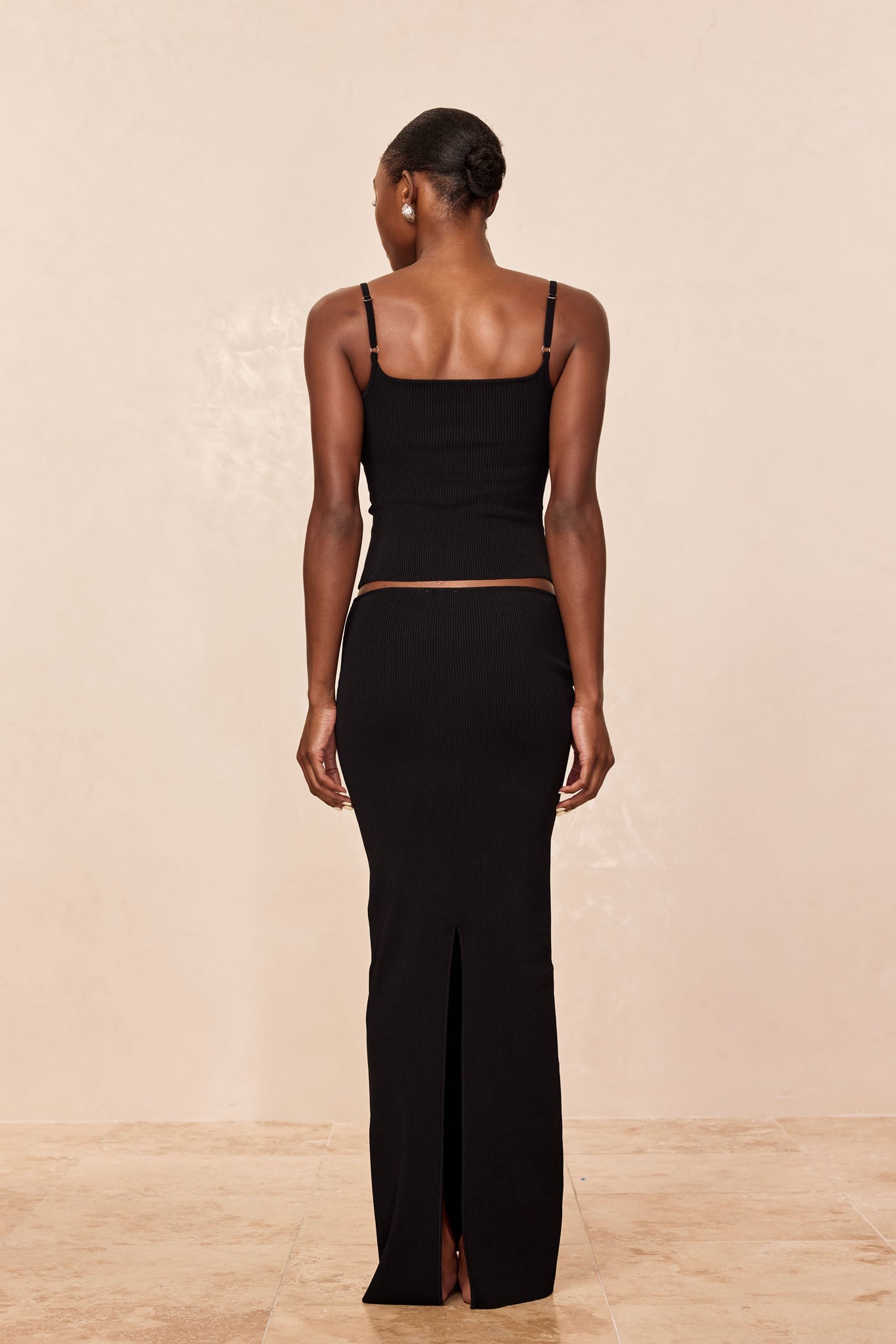 Woman shown from back wearing black ribbed camisole and matching long skirt with center back slit, hair in a low bun.