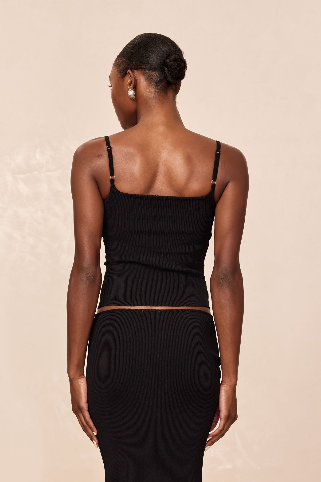Rear view of a woman with a low bun wearing a black ribbed tank top and matching skirt and gold hoop earrings.
