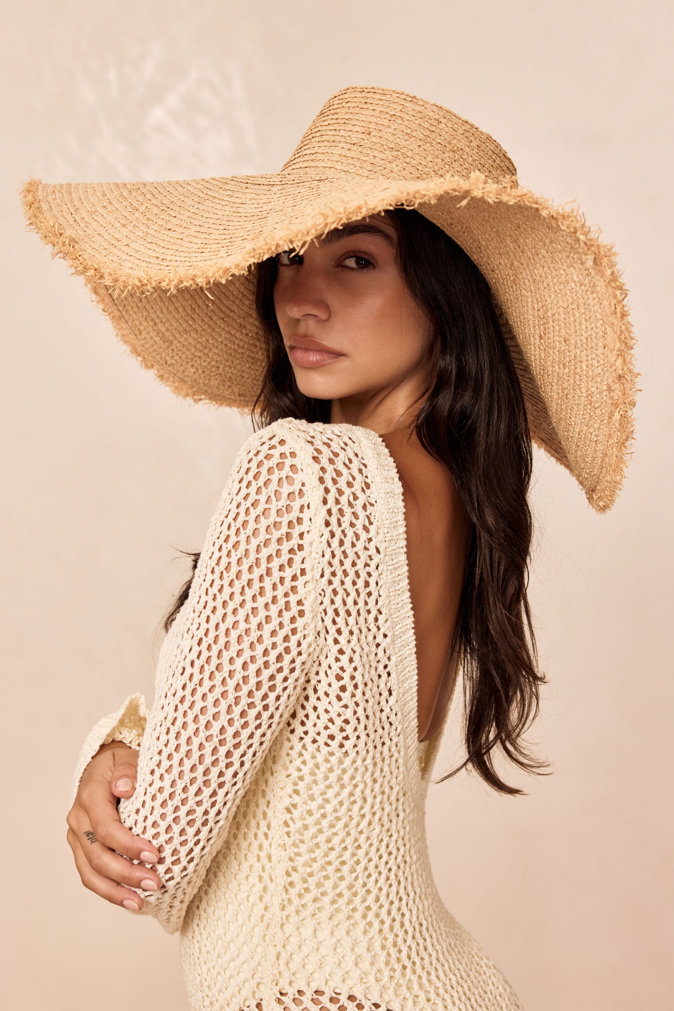 Woman wearing a wide straw sun hat and cream crocheted open-back sweater, glancing over her shoulder