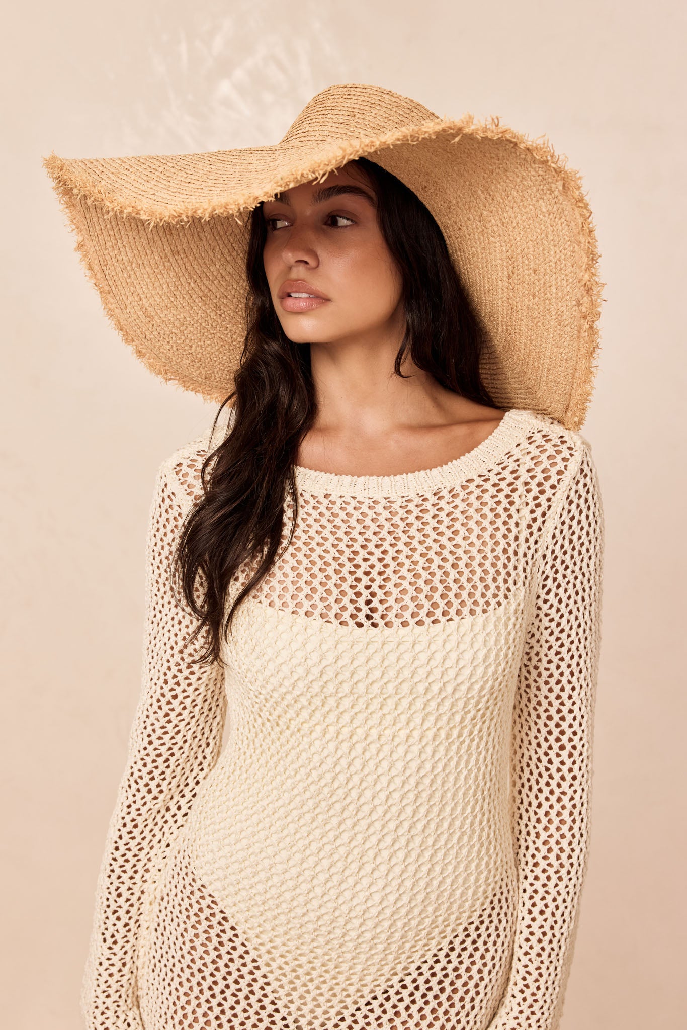Woman wearing a wide-brim straw sun hat and cream open-knit coverup over a swimsuit, gazing left.
