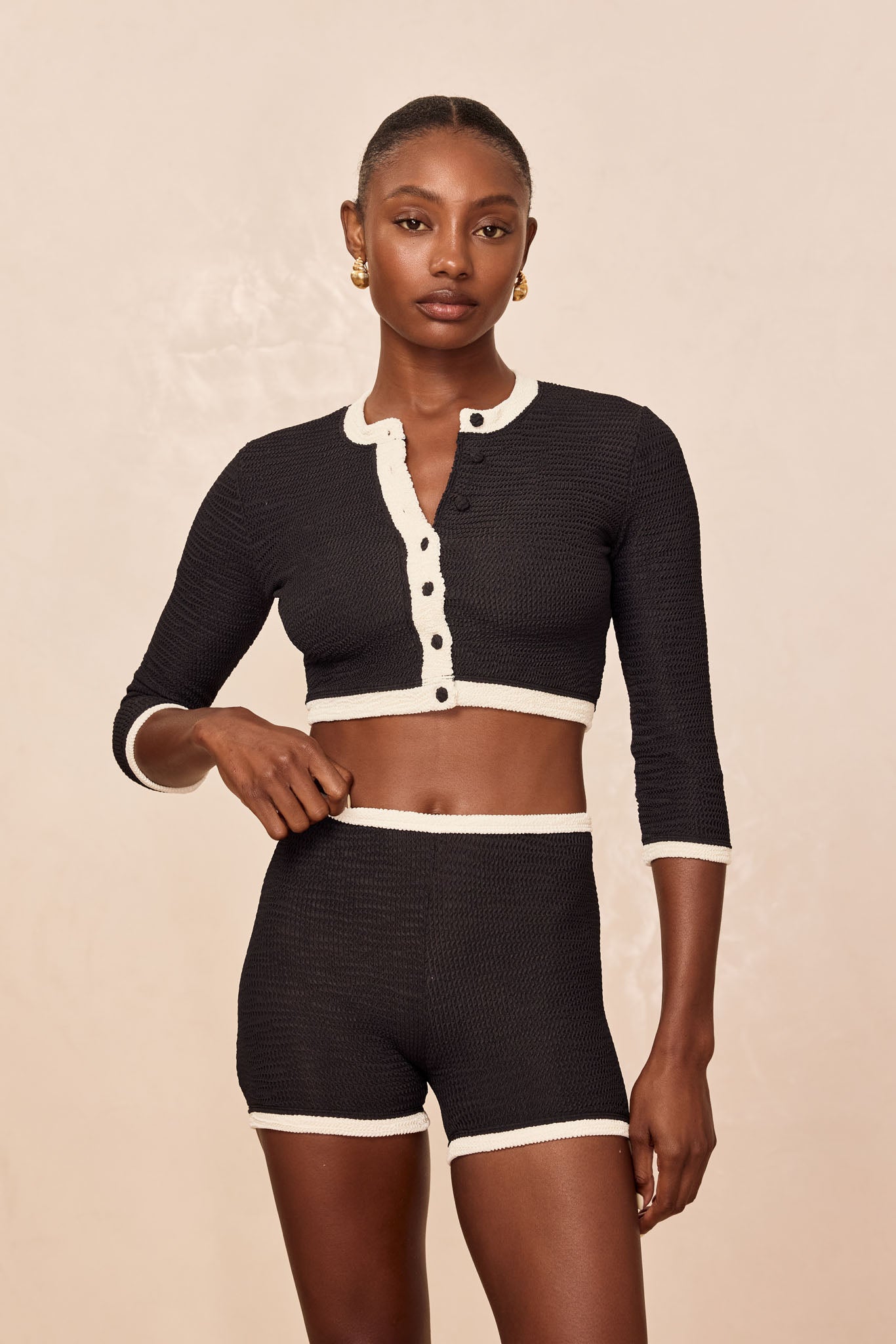 Woman modeling black textured knit cropped cardigan with white trim and matching high-waist shorts against a neutral backdrop.