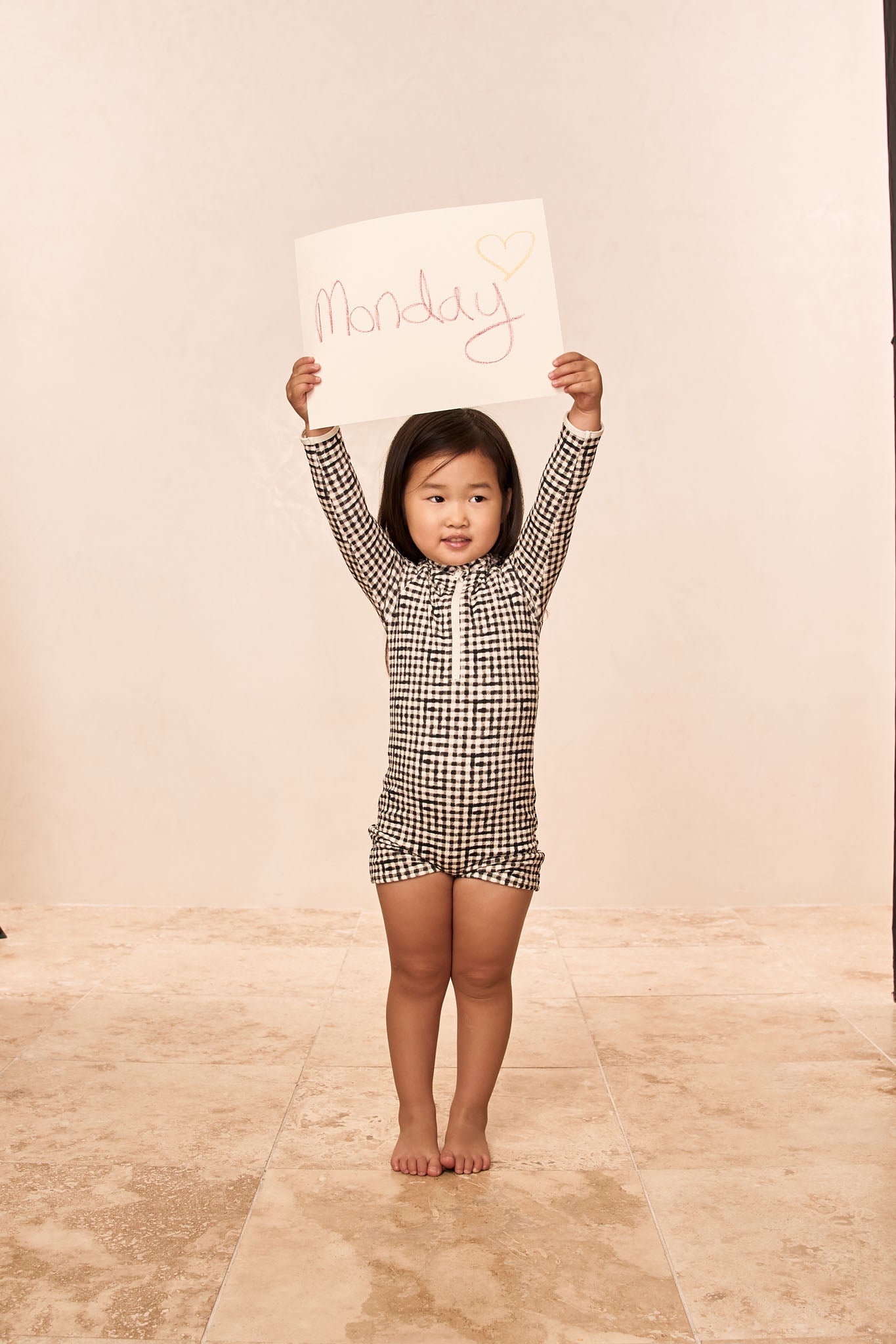 Young girl in black-and-white check romper standing barefoot on tile, holding a sign that reads 'Monday' with a drawn heart.