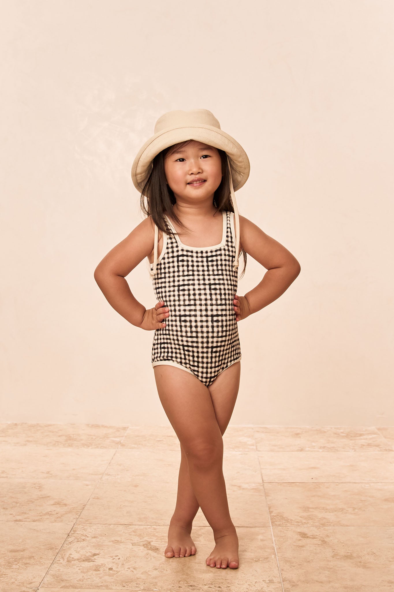 Young girl wearing checked swimsuit and beige bucket hat, standing barefoot on tile floor, hands on hips, smiling.