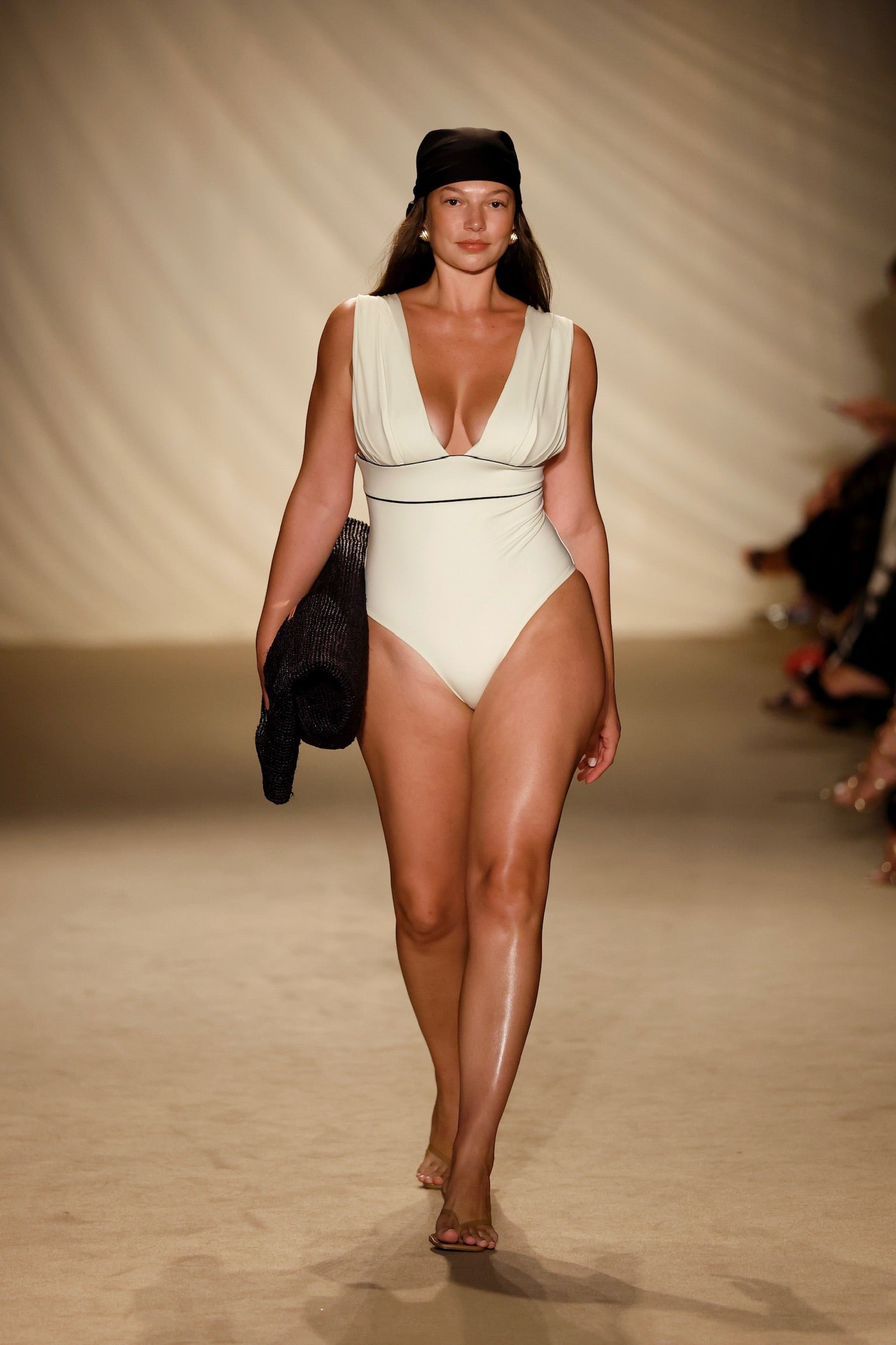 Runway model wearing a cream one-piece swimsuit (deep V), black headscarf, holding a rolled black knit cover-up.