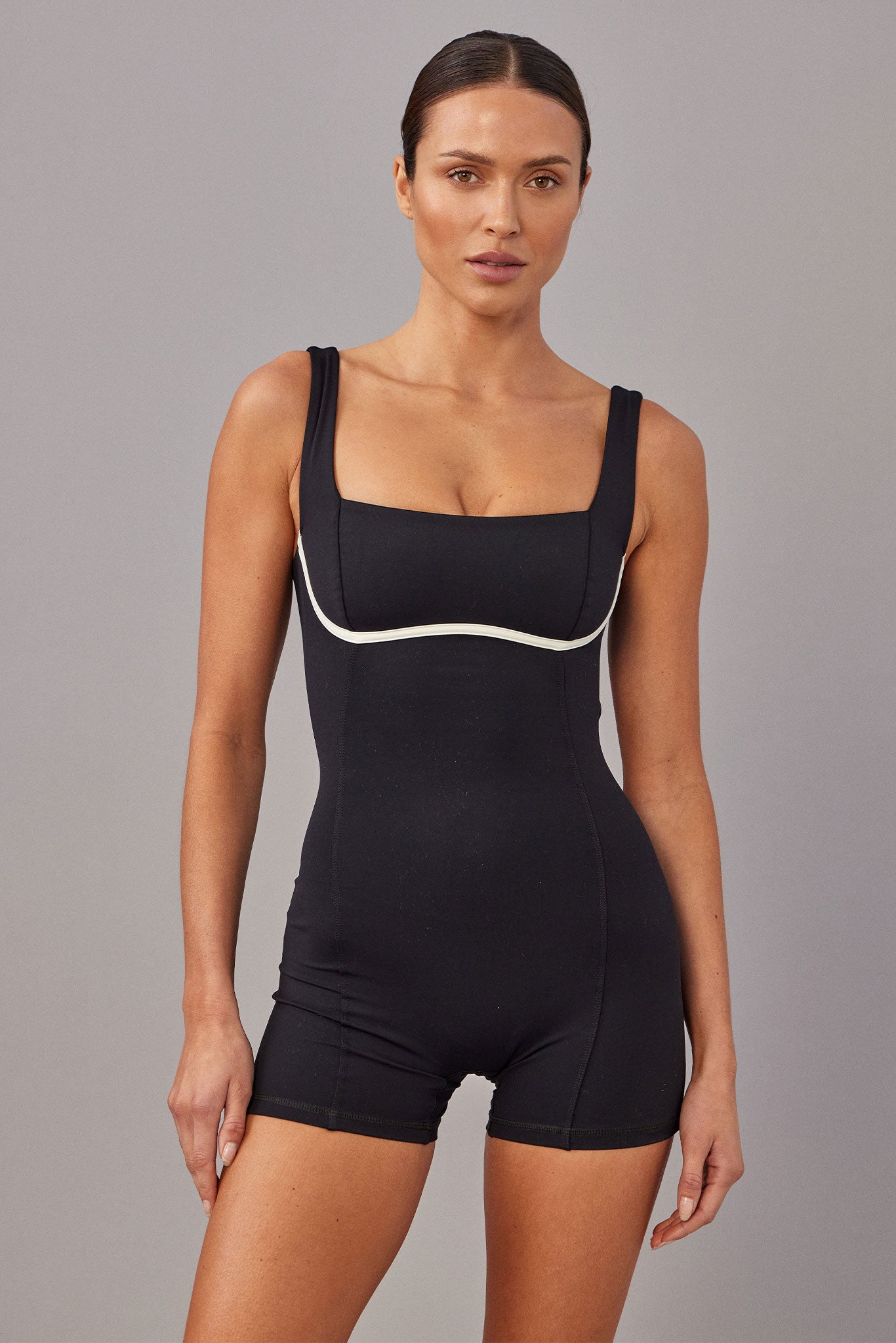 Model wearing a black fitted romper with square neckline and white piping under the bust, hands at sides