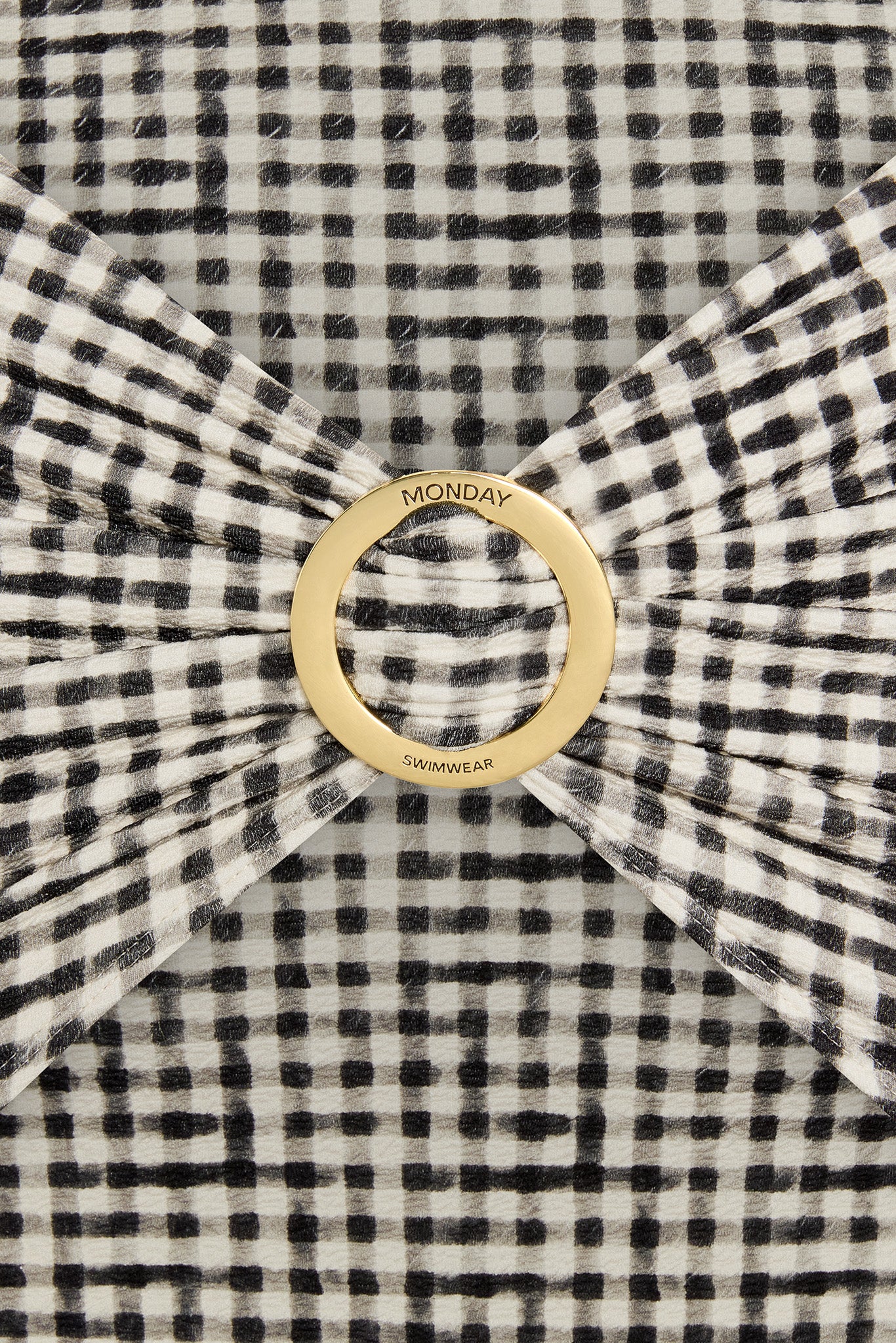 Black-and-white gingham fabric gathered through a gold ring engraved 'MONDAY' and 'SWIMWEAR'.