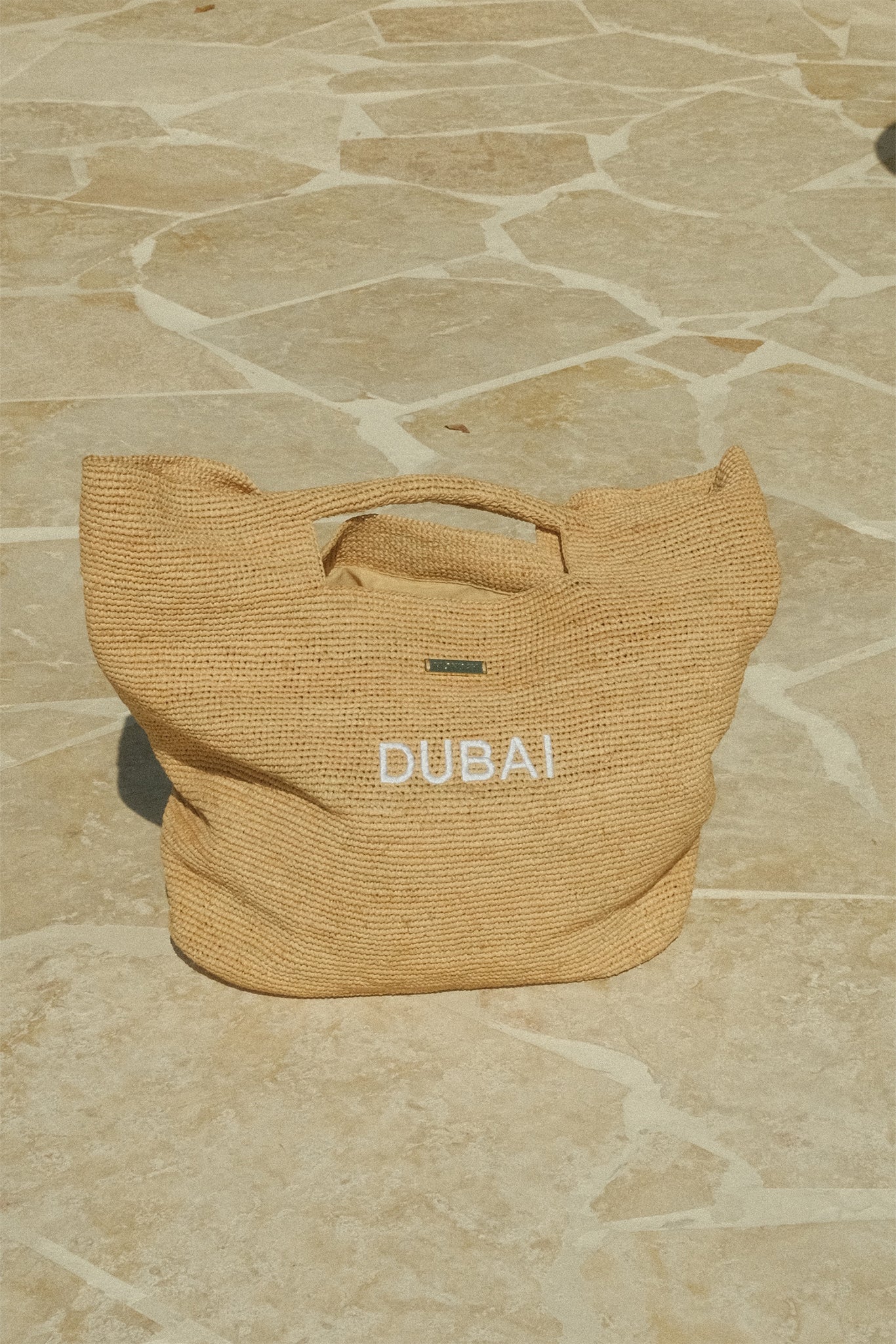 Woven straw tote bag on stone tiles with white embroidered text 'DUBAI'.