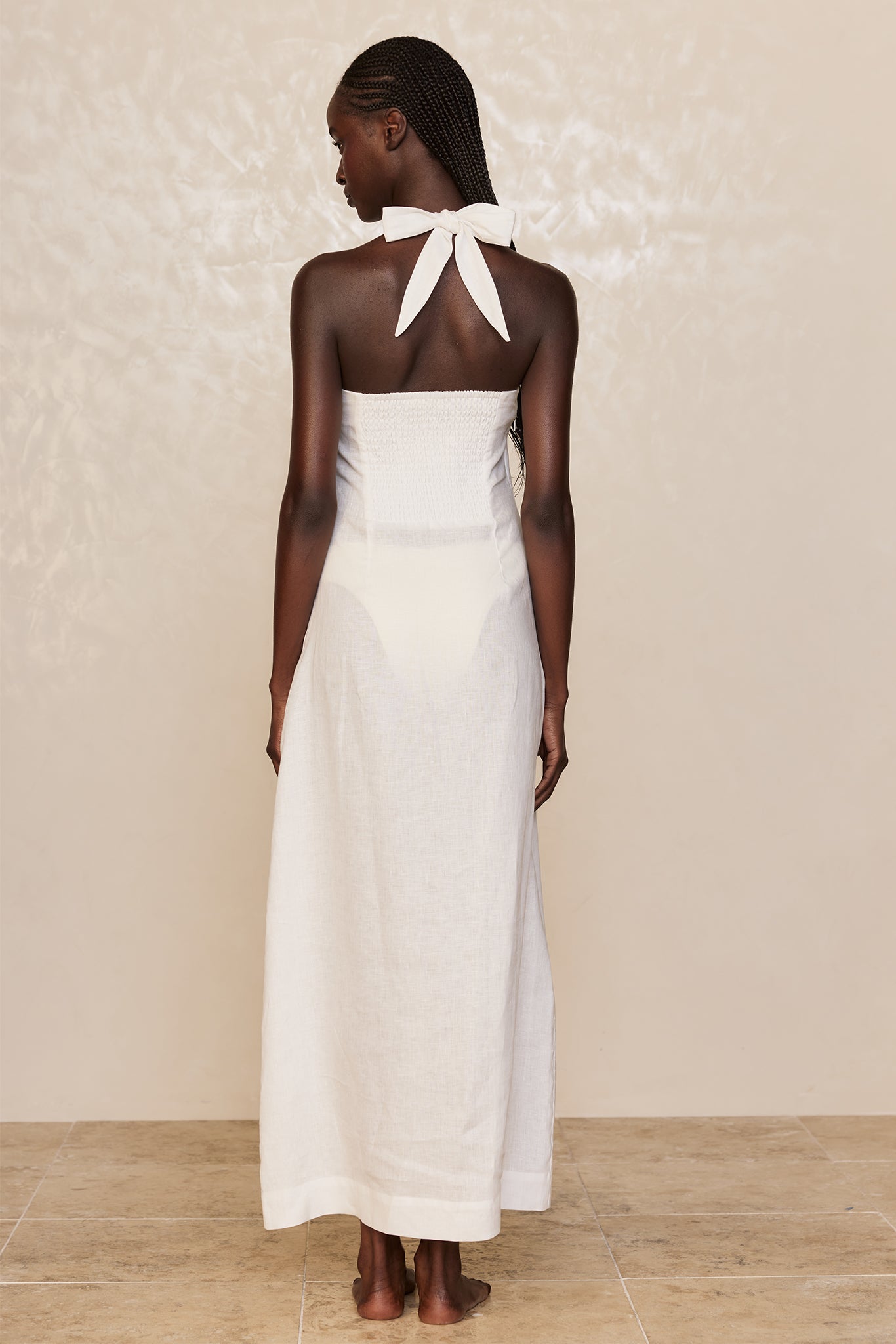 Model wearing a white strapless linen halter dress seen from the back; tied bow at the nape and floor-length skirt.