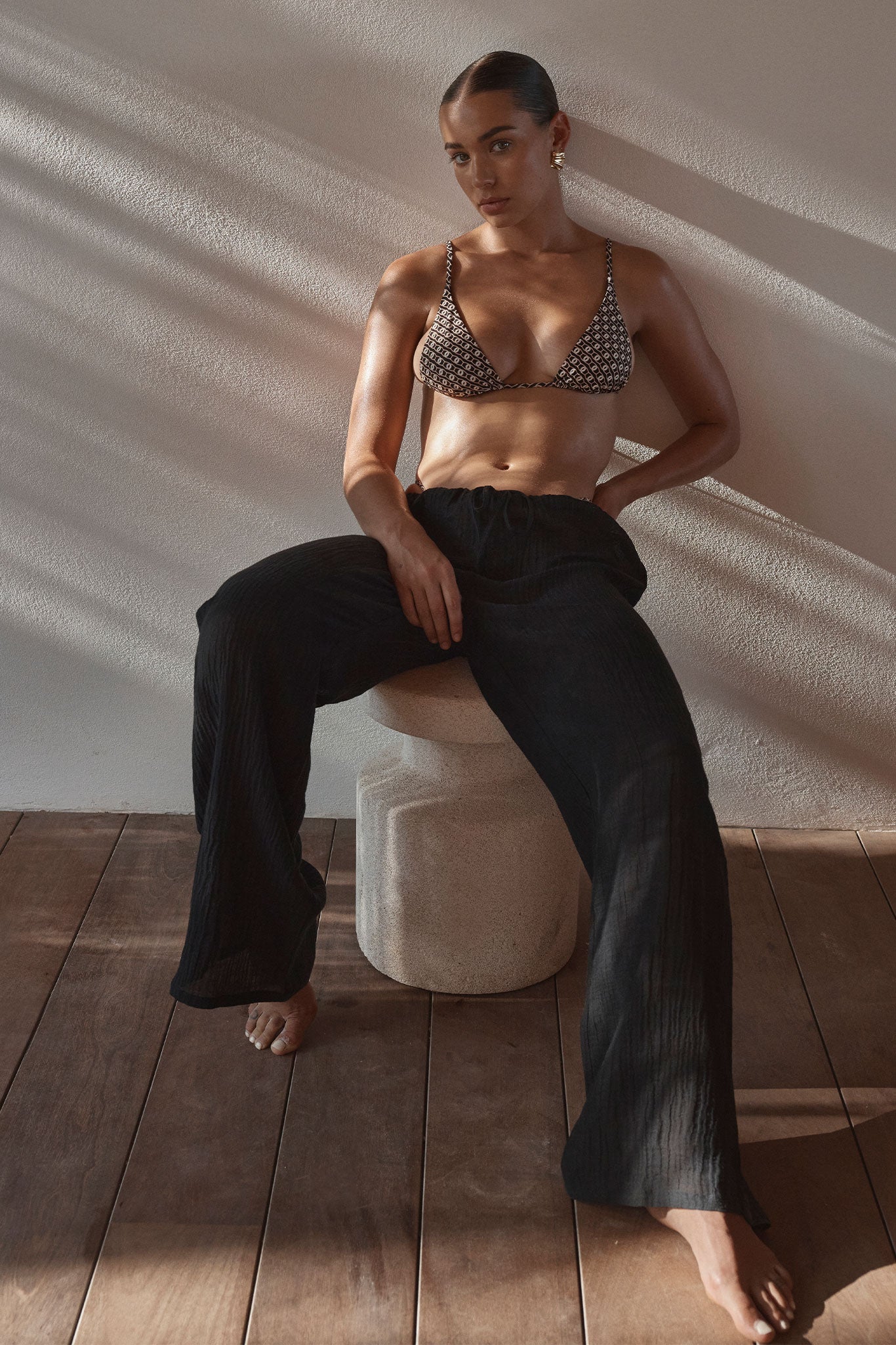 Woman seated on a stone stool wearing patterned triangle bikini top and loose black pants, sunlight casting shadows on wall