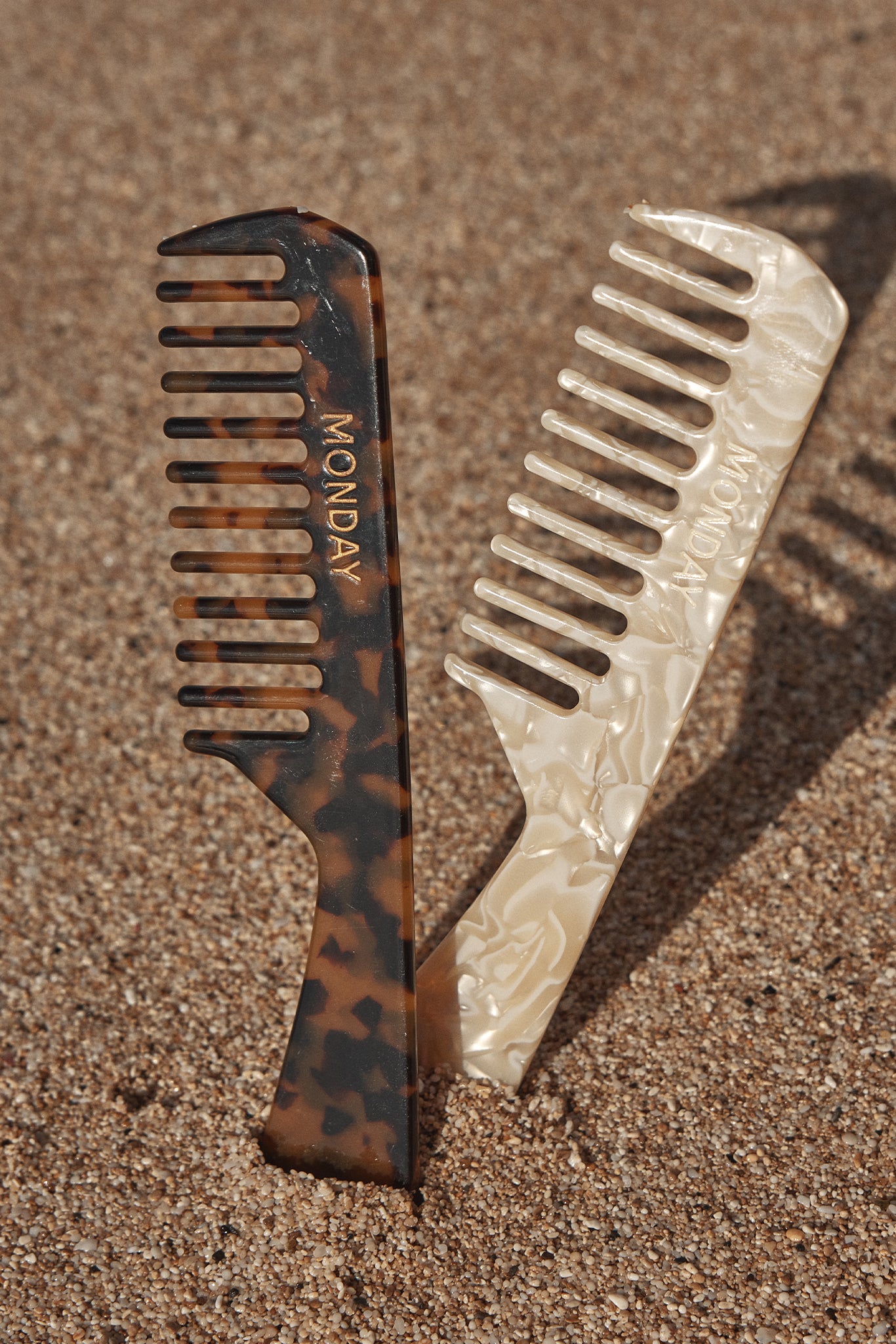 Two wide-tooth hair combs—tortoiseshell and pearlescent—standing in sand; both combs embossed with the word MONDAY.