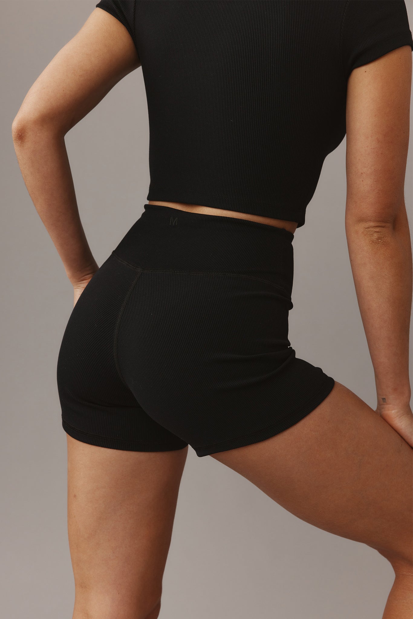 Rear view of model wearing black ribbed crop top and high-waist shorts; small 'M' on waistband and tattoo 'III' on wrist.