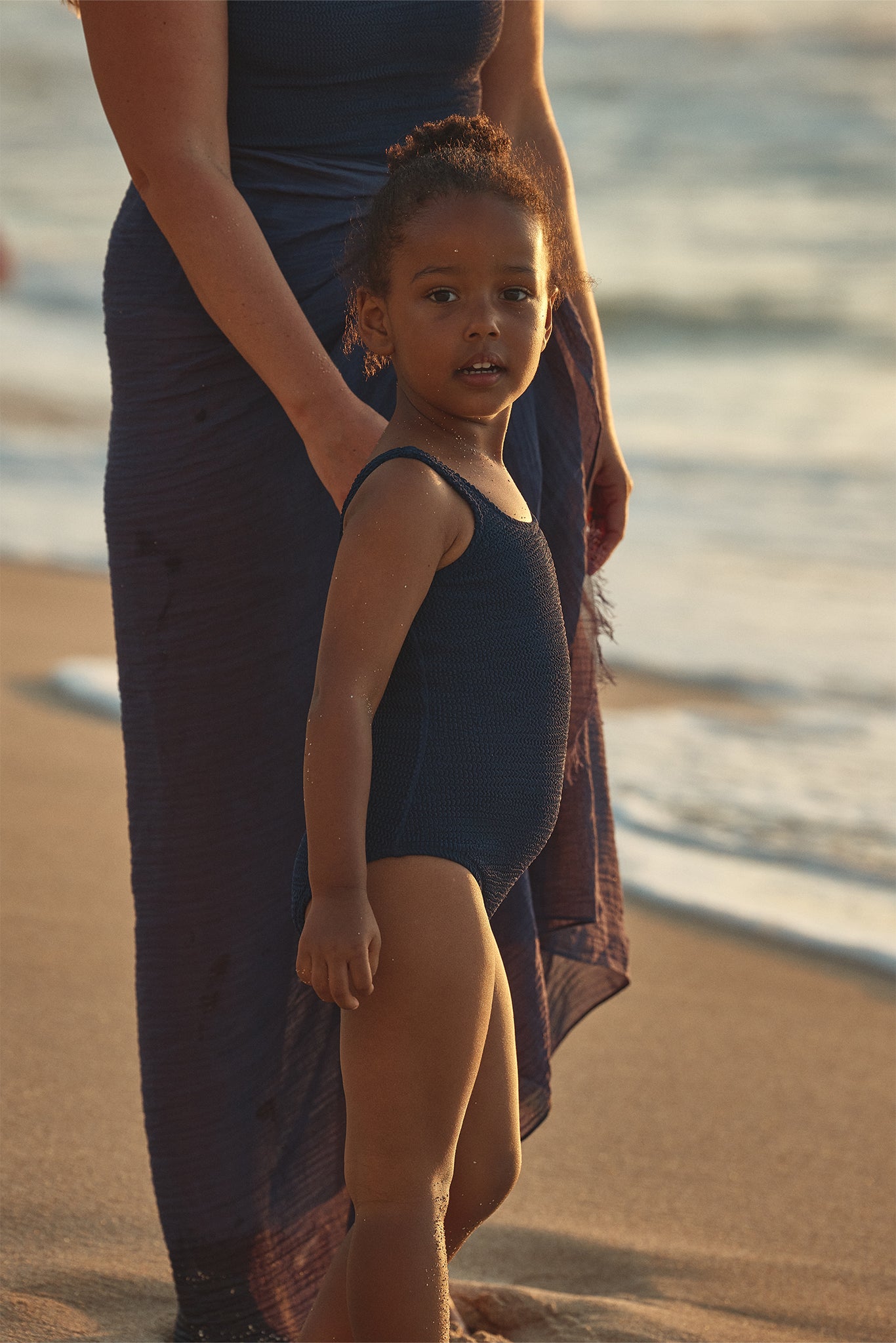 Young child in a navy swimsuit holding an adult's hand on a sandy beach at sunset