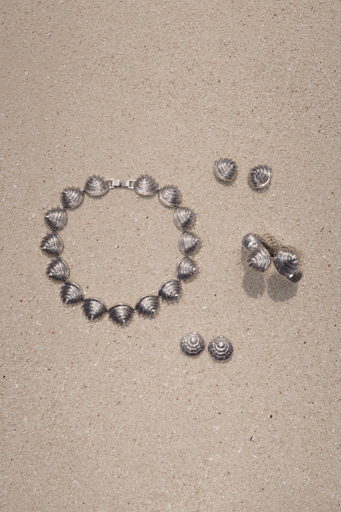 Silver seashell-style necklace with three pairs of matching silver shell earrings arranged on sand