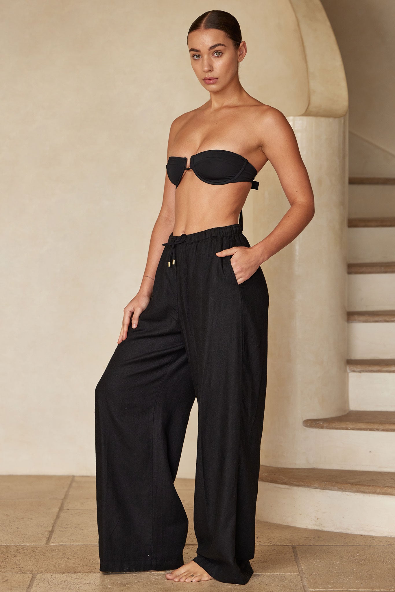Model wearing black bandeau top and wide-leg black linen pants, standing barefoot beside curved staircase in neutral room.