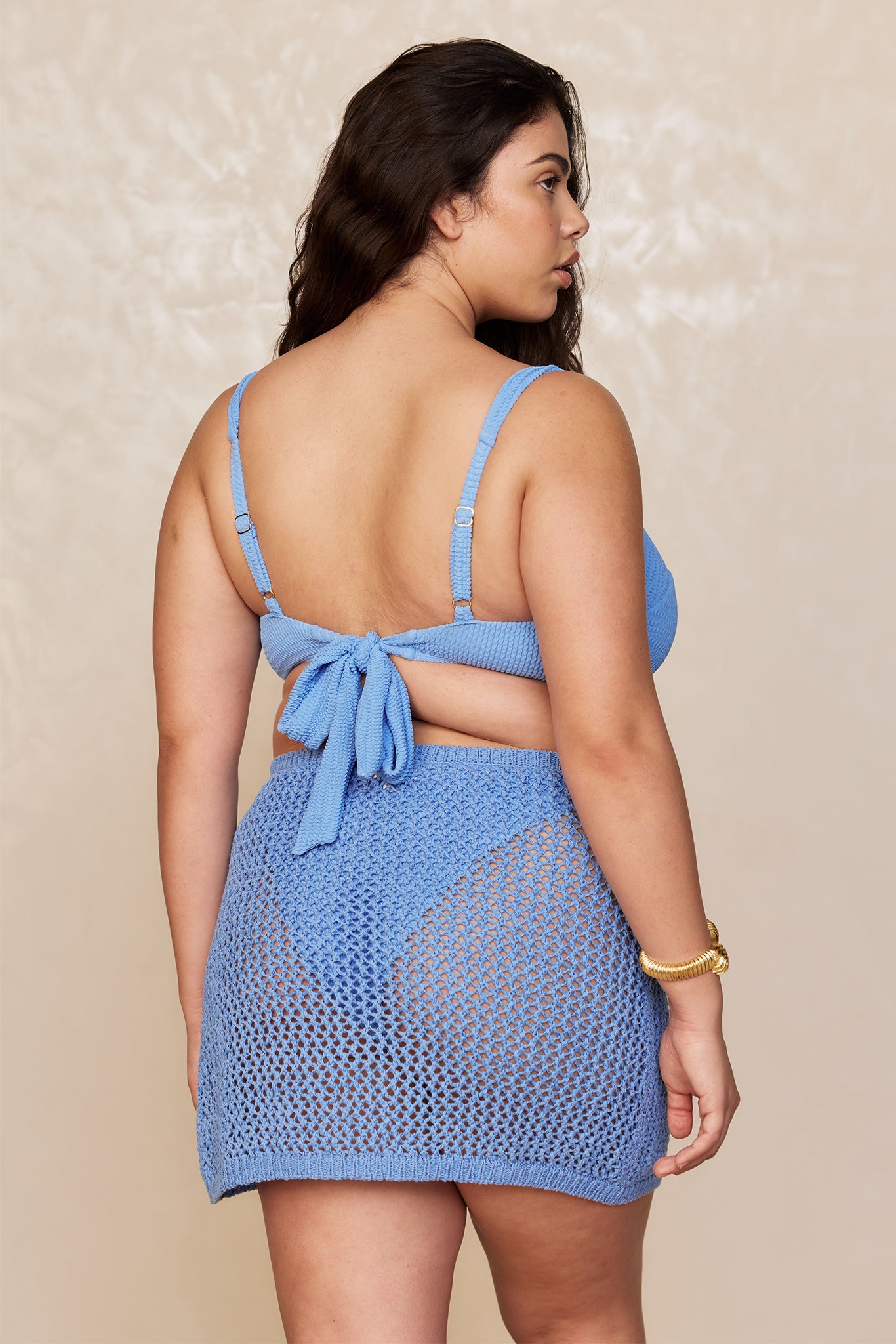 Woman seen from back wearing light blue knit bikini top tied at back and a matching crochet mini skirt with a gold bracelet