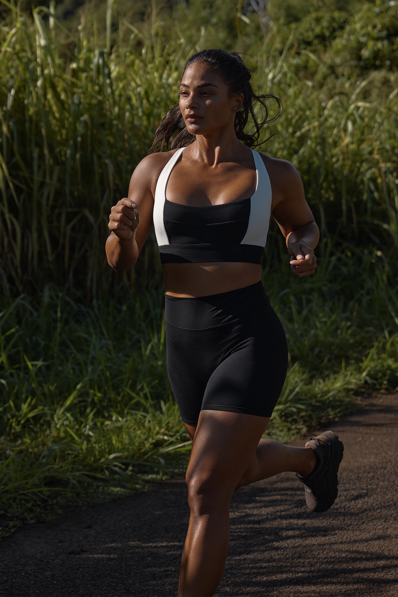 Athletic woman running on a sunlit paved path beside tall grass, wearing black shorts and black sports bra with white straps.