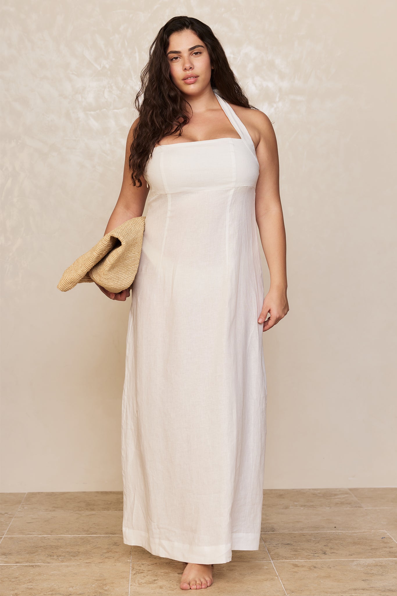 Woman in white halter maxi linen dress, barefoot on tiled floor, holding a folded straw sunhat at her side.