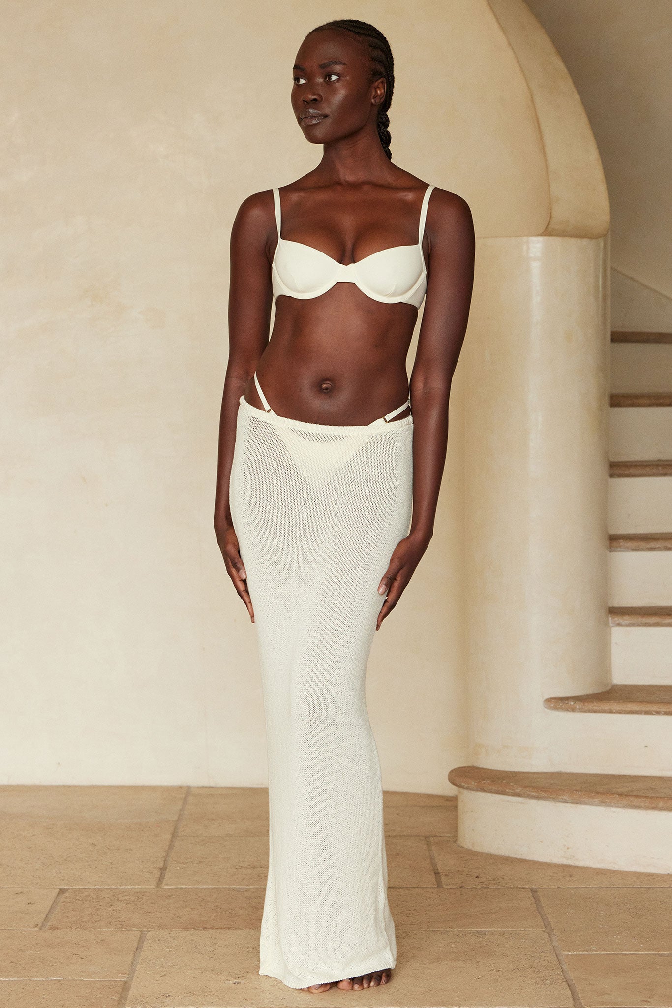 Model wearing cream underwire bra and sheer knit cream maxi skirt, standing barefoot by curved stone staircase.