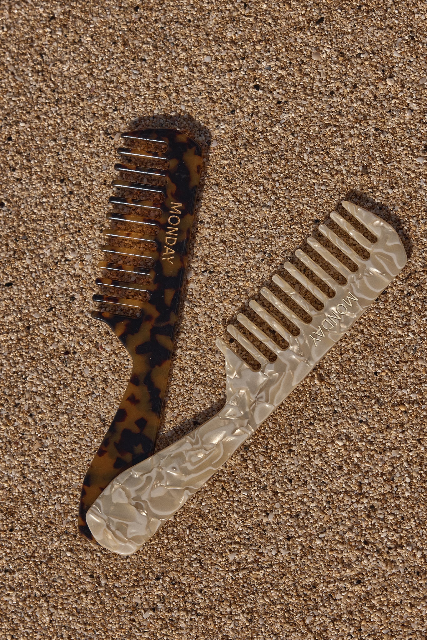 Two wide-tooth combs on sand: tortoiseshell and pearlescent styles, each embossed with the word MONDAY.