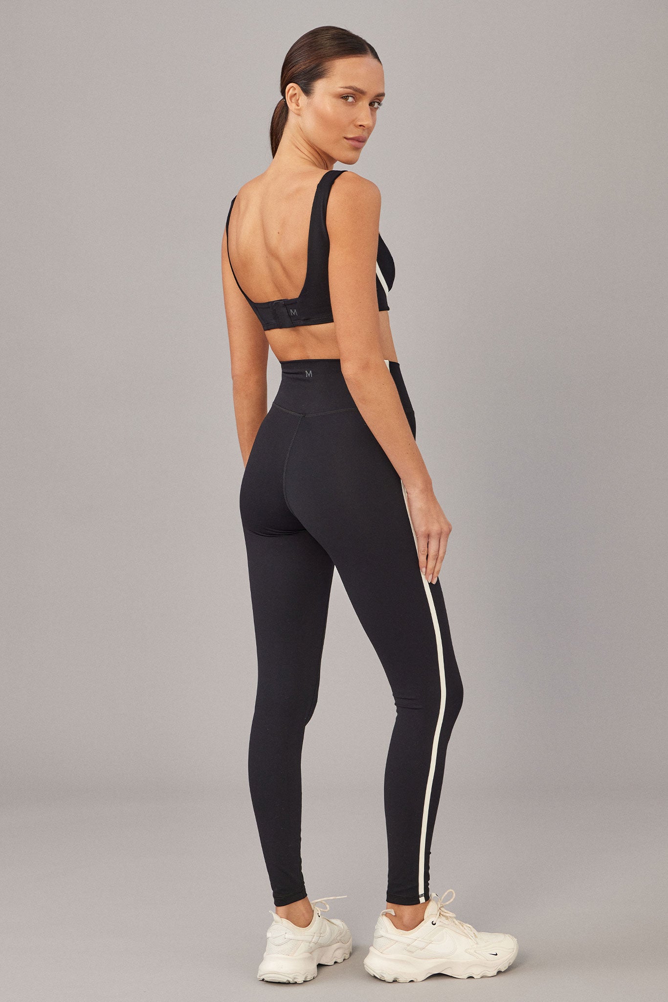 Woman wearing black high‑waist leggings with white side stripe and matching sports bra; 'M' logo visible on waistband.