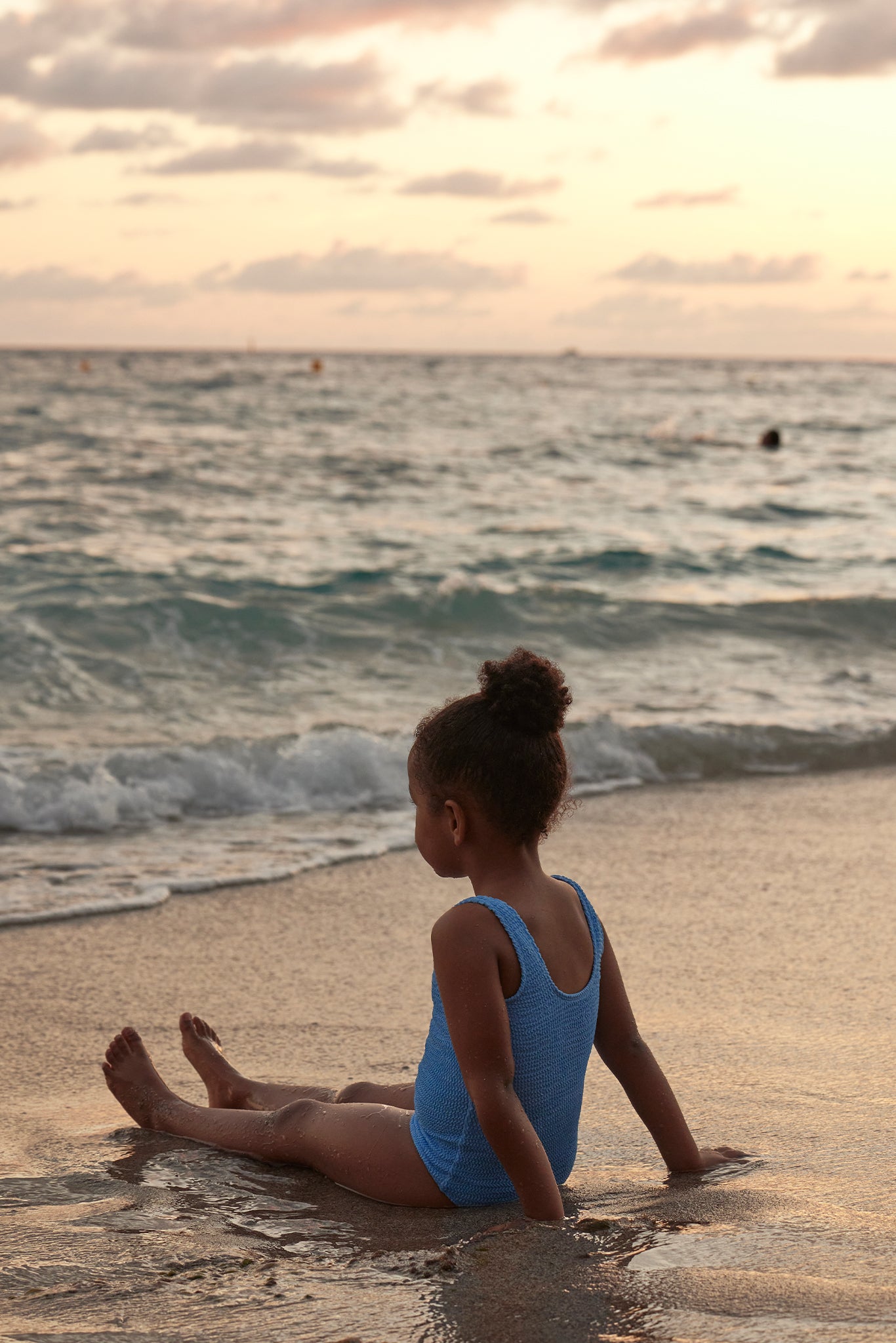 Child in a blue swimsuit sitting on wet sand at sunset, facing the ocean waves.