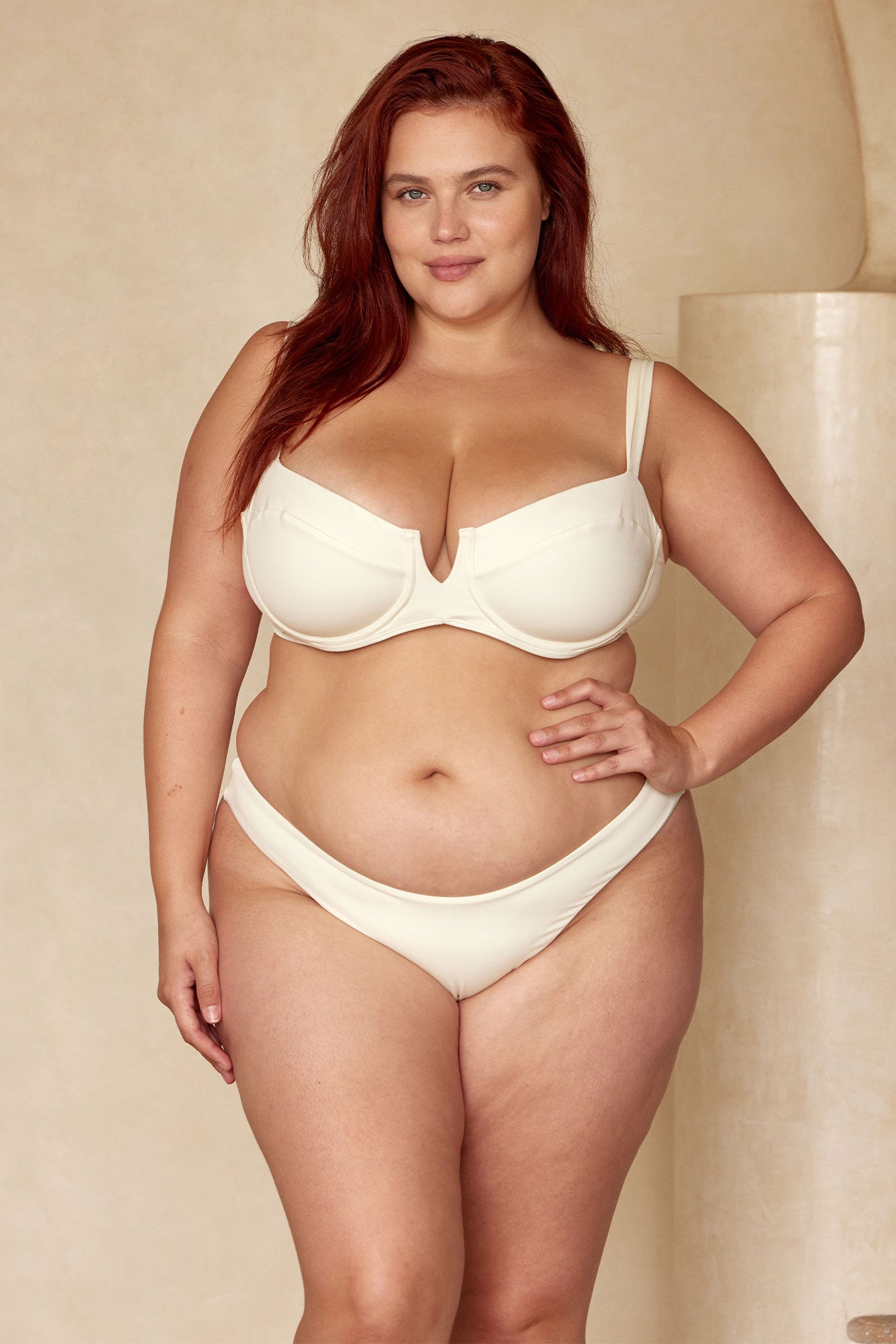 Plus-size woman standing in cream undergarments with hand on hip, neutral background, confident relaxed expression.