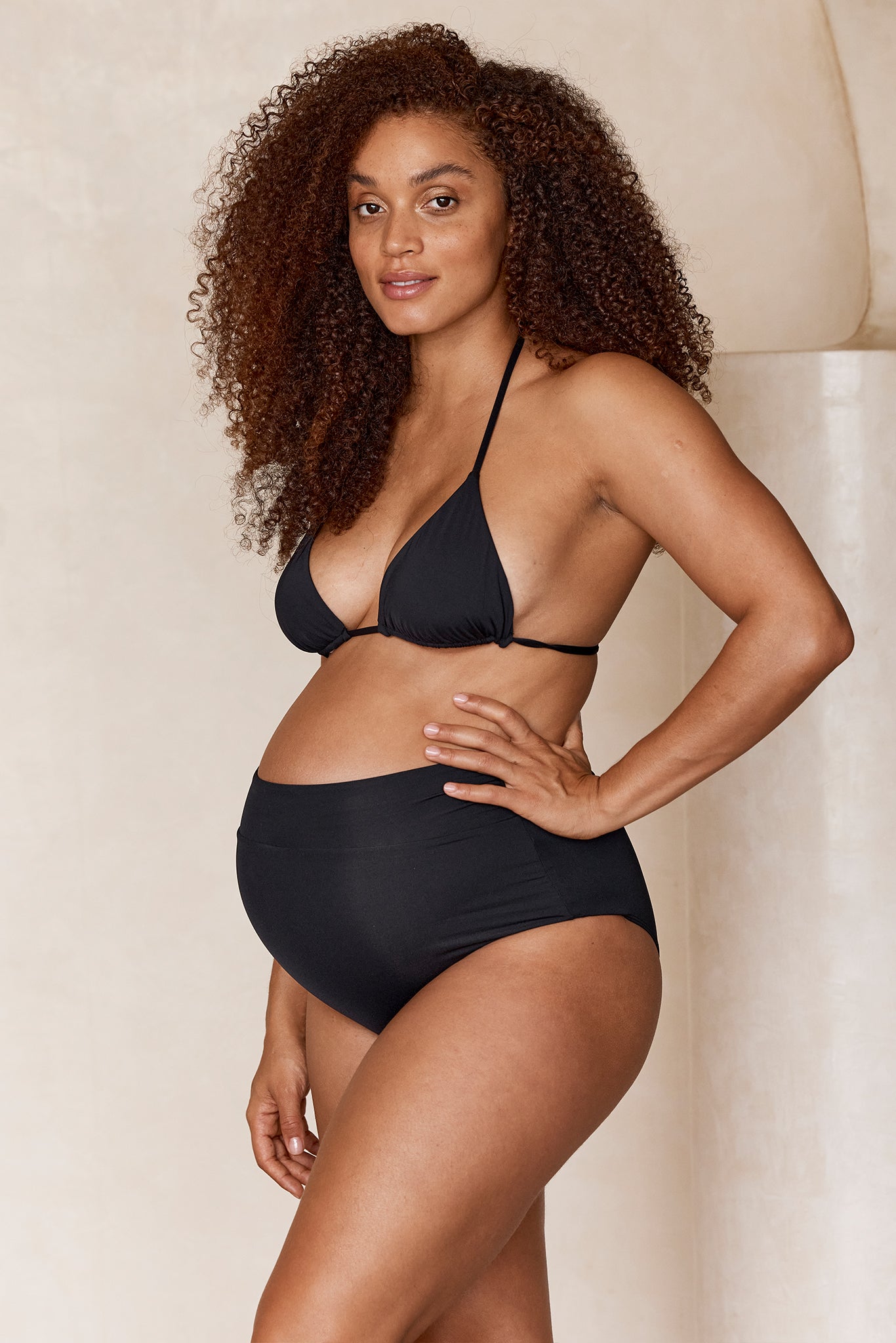 Pregnant woman standing in a black bikini top and high-waist swim bottoms, hand on hip, curly hair, neutral background