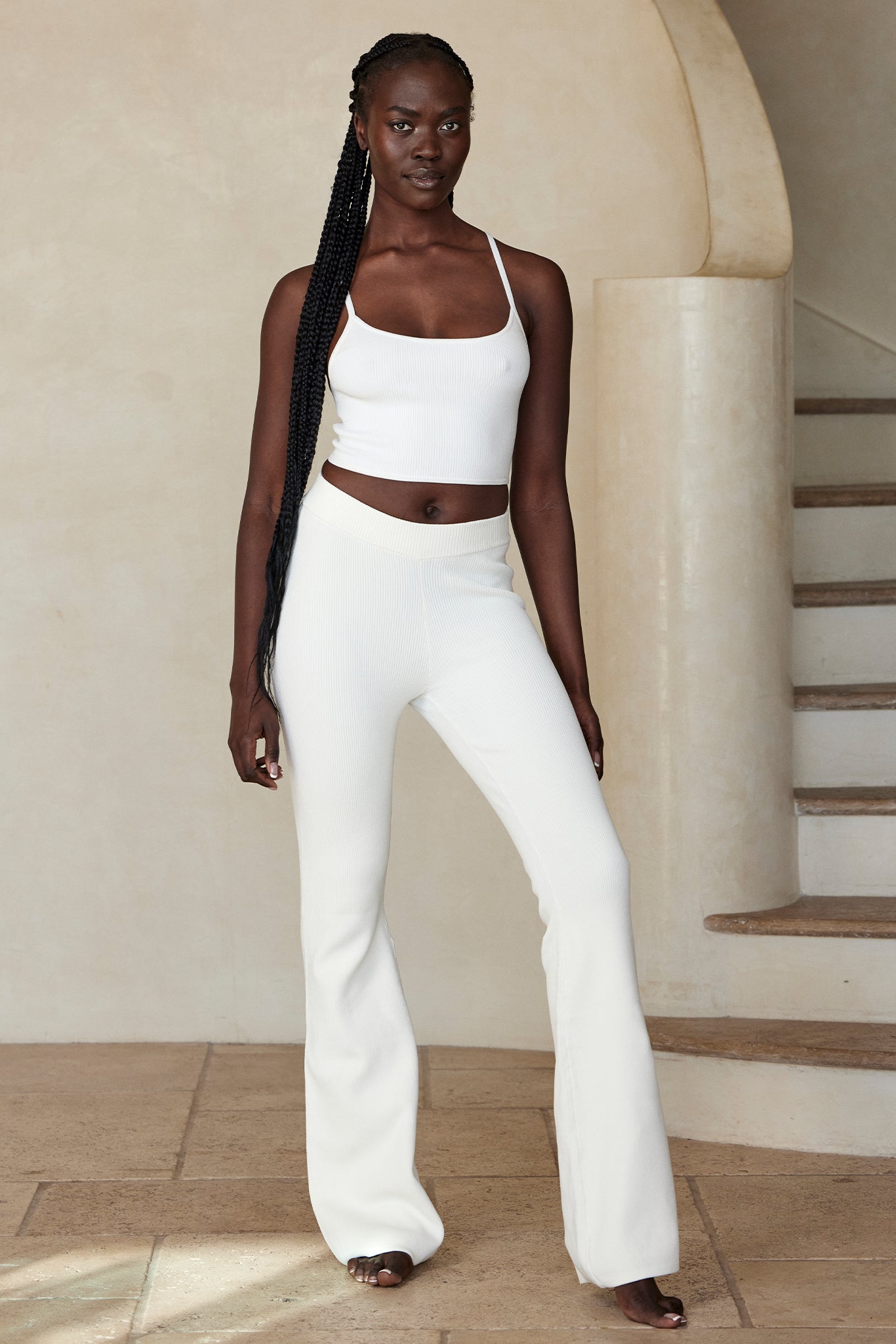 Woman with long braided hair wearing a white cropped tank and ribbed flared pants, barefoot, standing by a curved stone staircase.
