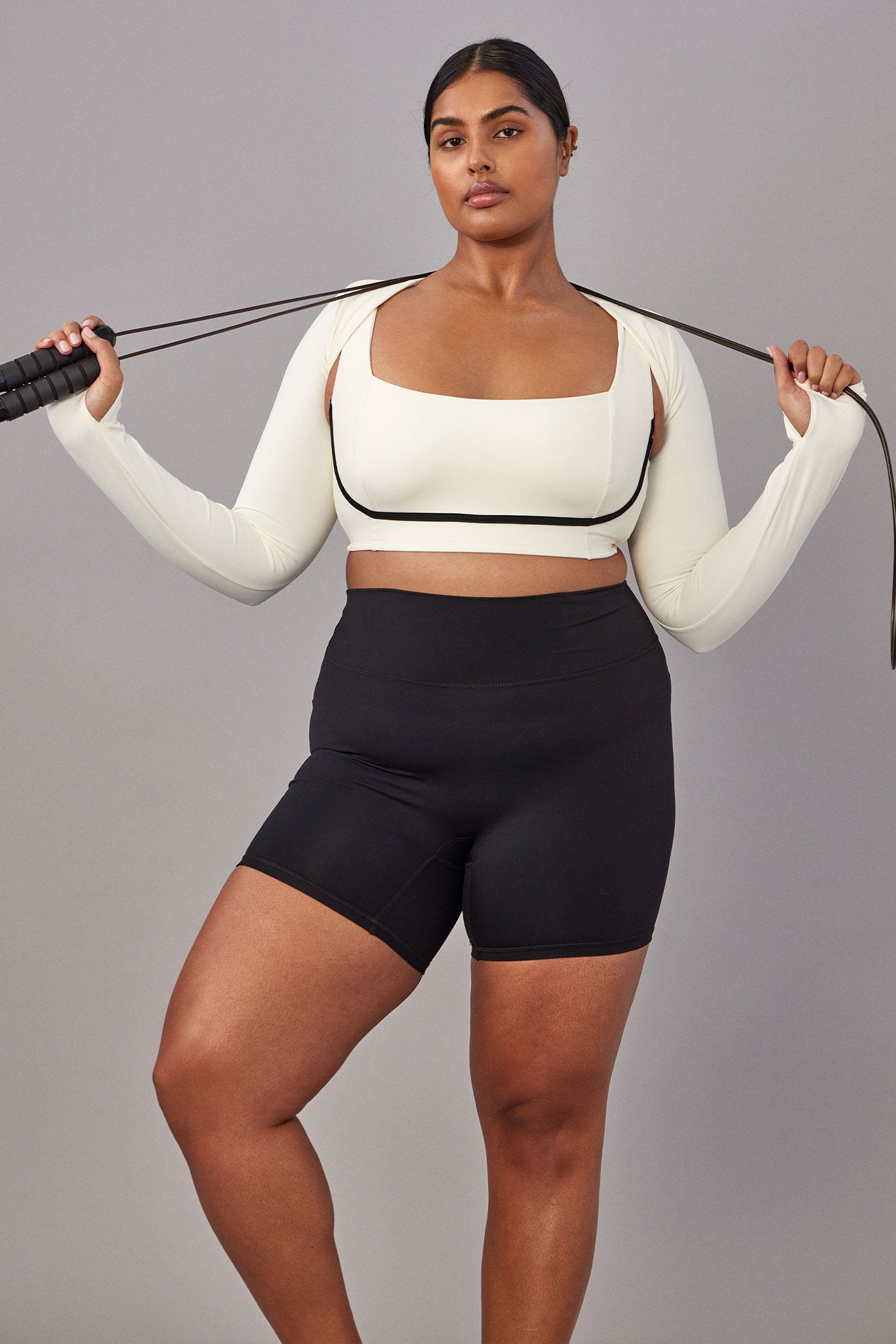 Plus-size woman in cream long-sleeve crop top and high-waist black bike shorts holding a jump rope behind her shoulders.
