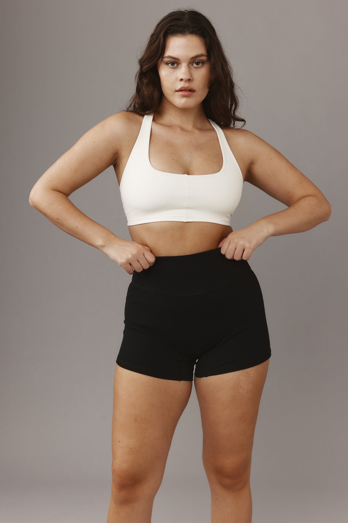 Woman adjusting the waistband of black high-waist shorts, wearing a white sports bra, facing the camera.