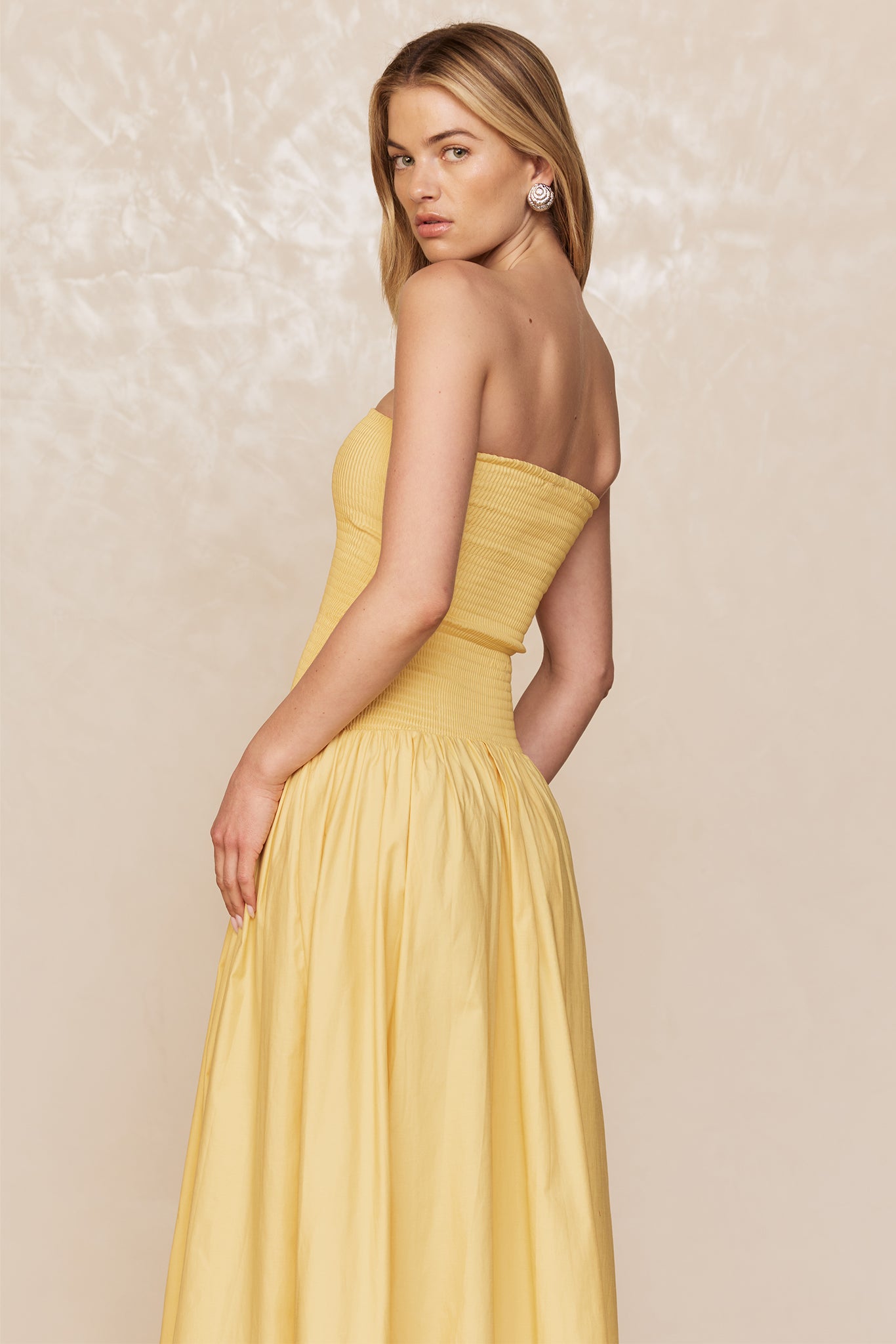 Model turned over her left shoulder wearing a strapless yellow maxi dress and spiral earring against a neutral textured background.