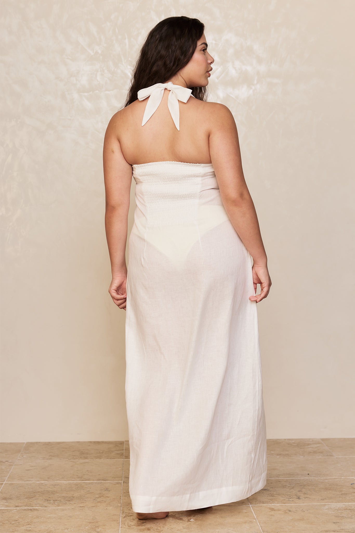 Back view of a woman wearing a strapless white linen maxi dress with a tied halter bow at the neck