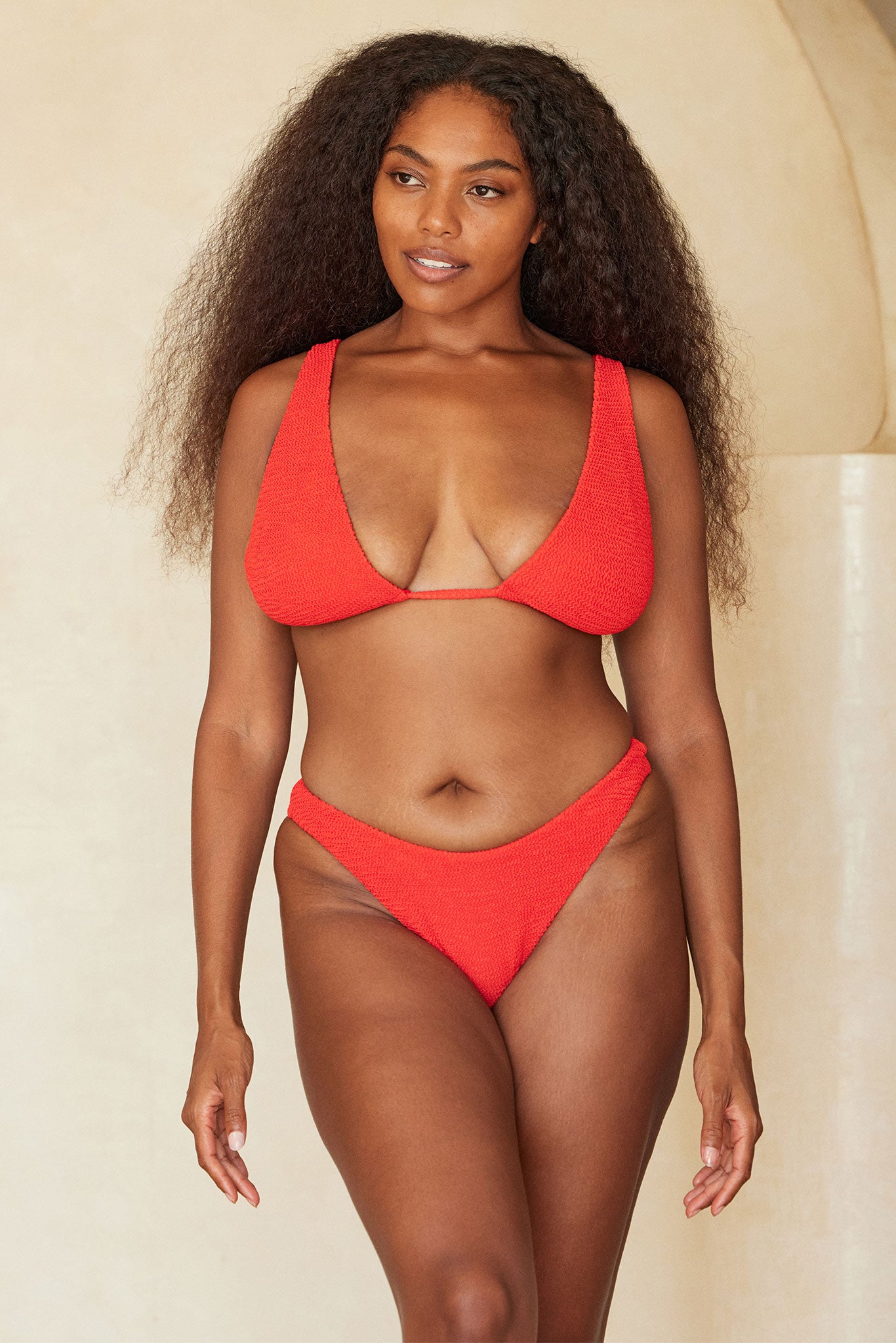 Model with long textured hair wearing a red textured bikini, standing against a beige background in a relaxed, three-quarter pose.