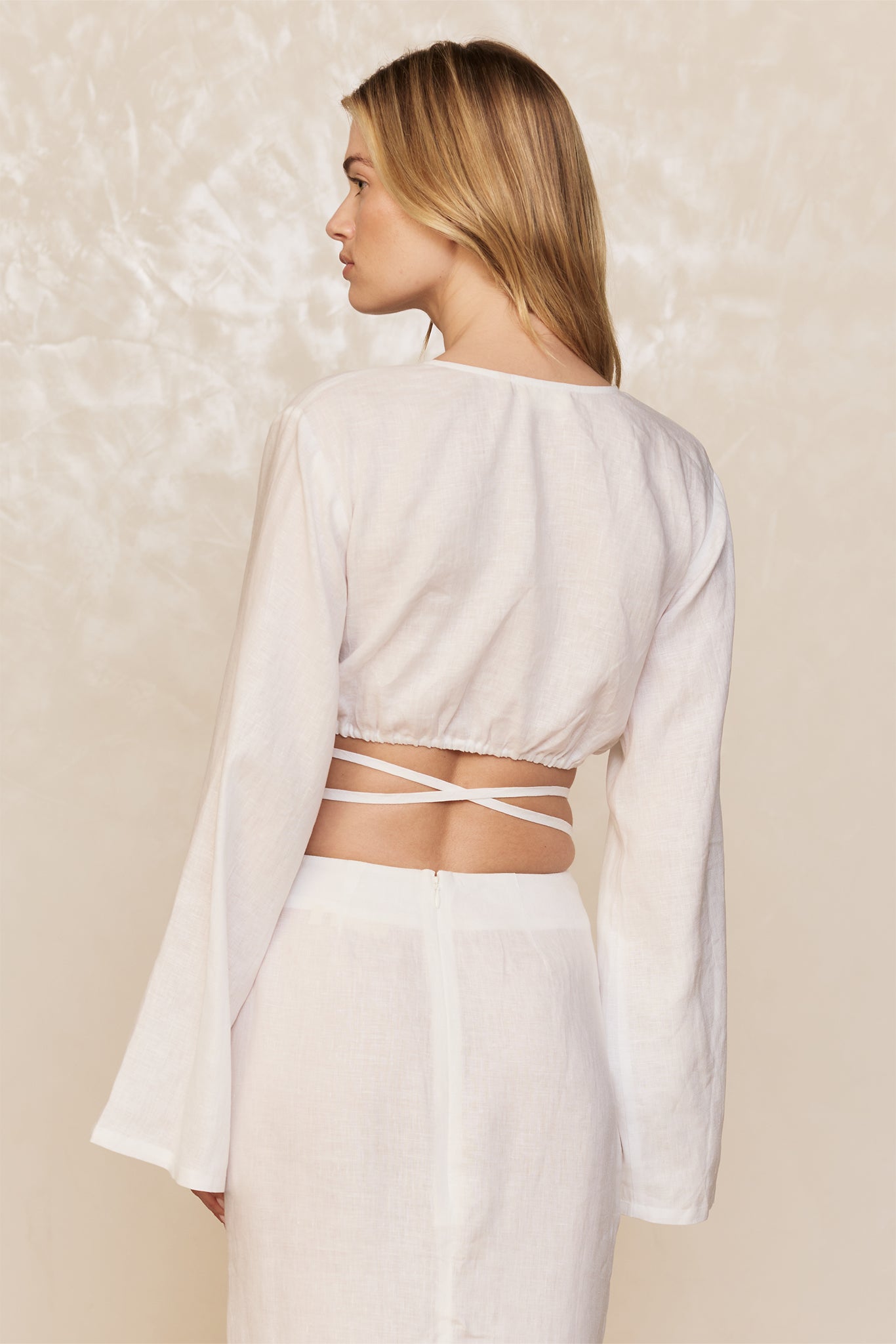 Woman viewed from back wearing a white long-sleeve cropped top with crisscross wrap straps at the waist and a white skirt