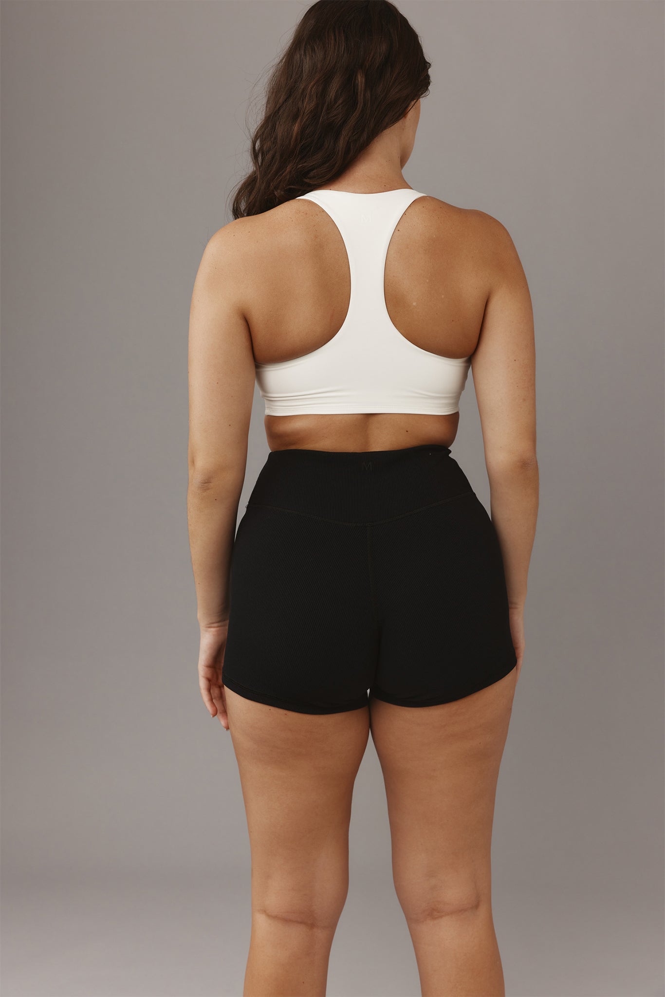 Back view of a model wearing a white racerback sports bra and high-waist black athletic shorts on a gray background.