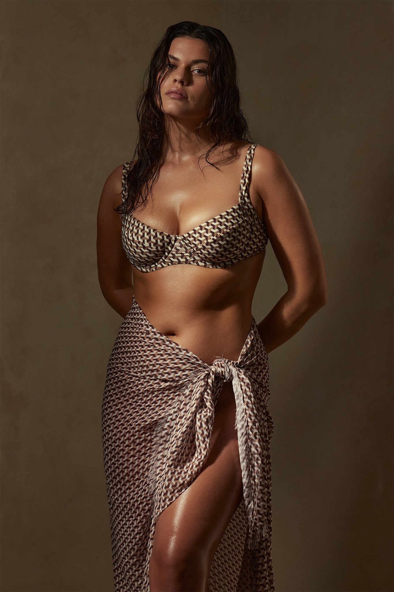 Woman wearing geometric-patterned bikini top and matching sarong tied at the hip, wet hair, neutral studio backdrop