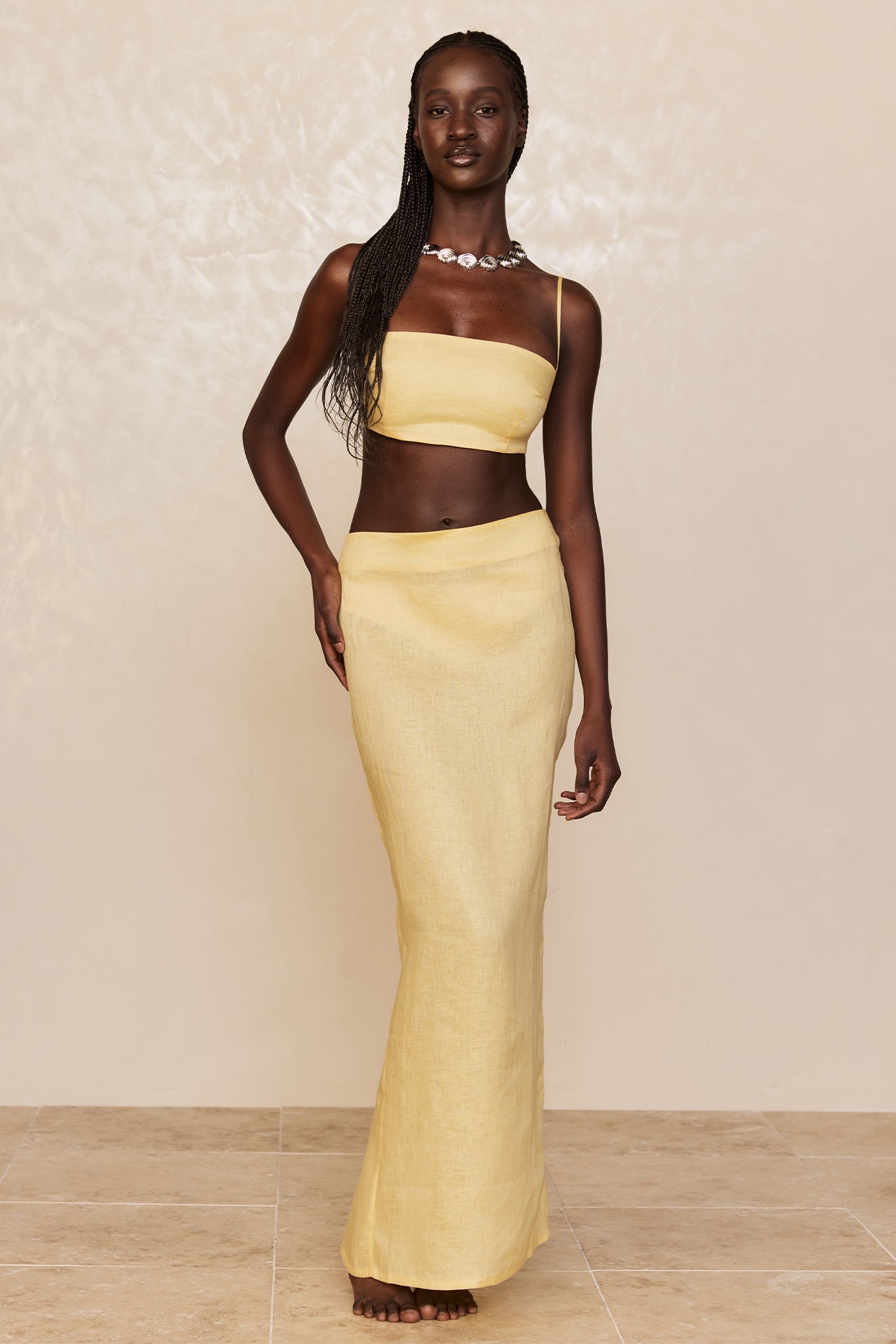Dark-skinned model wearing a yellow linen bandeau crop top with one spaghetti strap and a matching high-waisted maxi skirt, barefoot, neutral background.