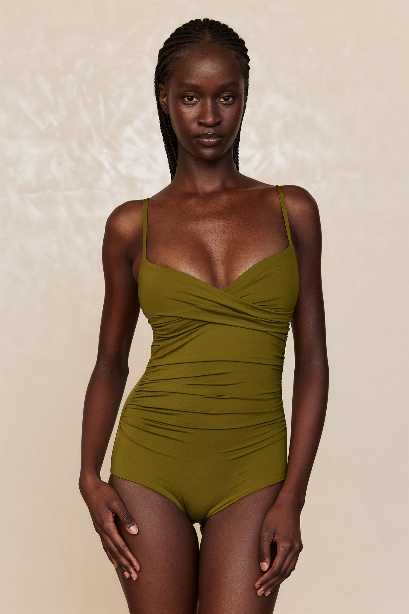 Dark-skinned woman modeling olive-green ruched one-piece swimsuit with thin straps, front view on neutral backdrop.