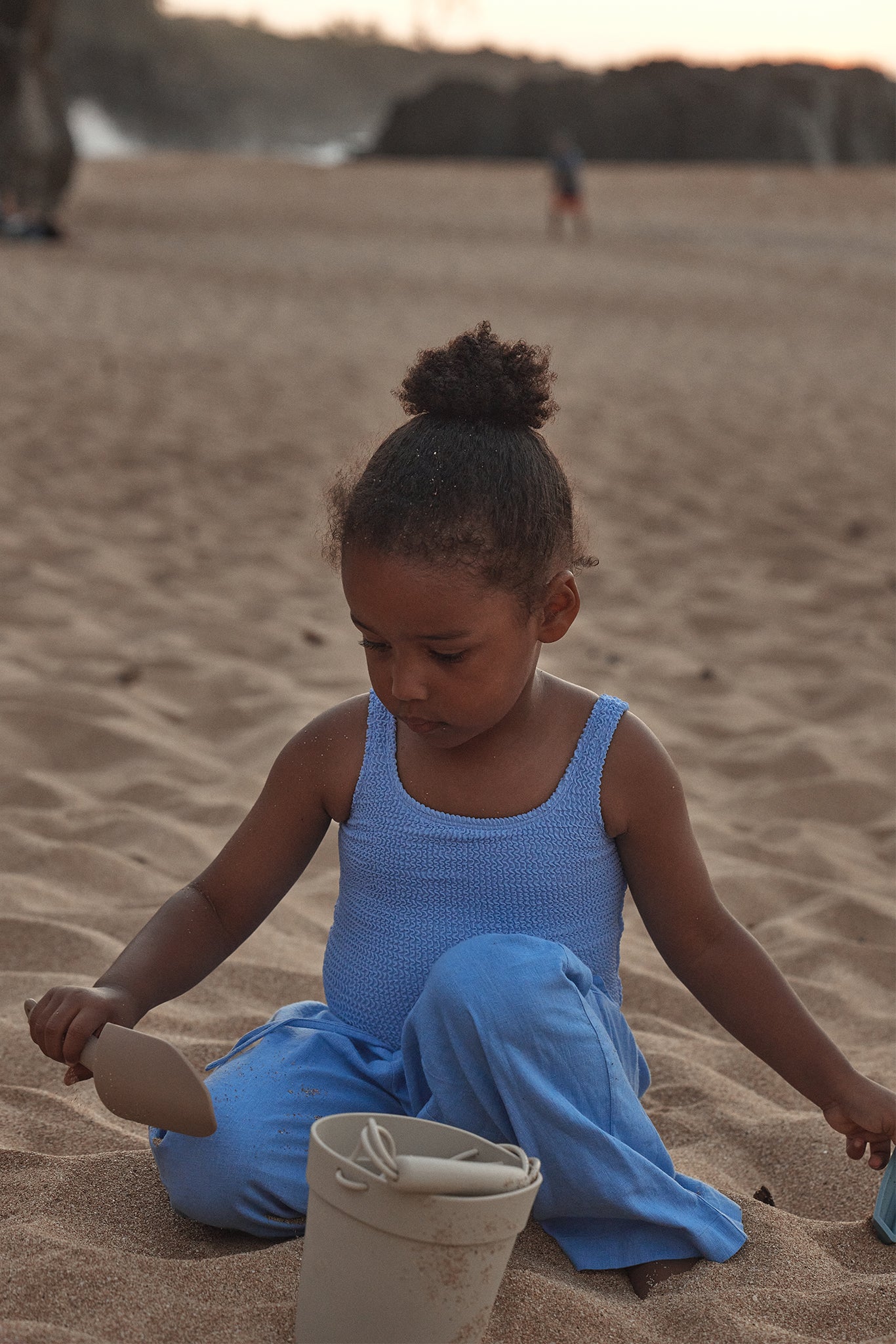 Young child in blue knit top and pants sitting on a sandy beach, using a small shovel with a bucket nearby.