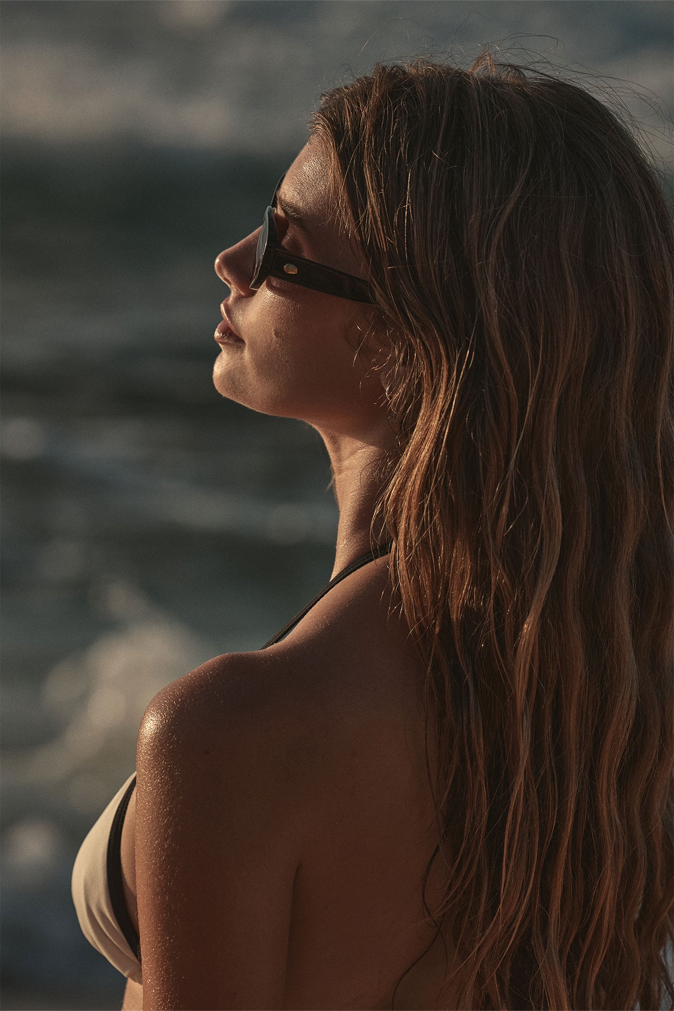 Young woman in sunglasses facing left on a beach at sunset, long wavy hair and sunlit shoulder.