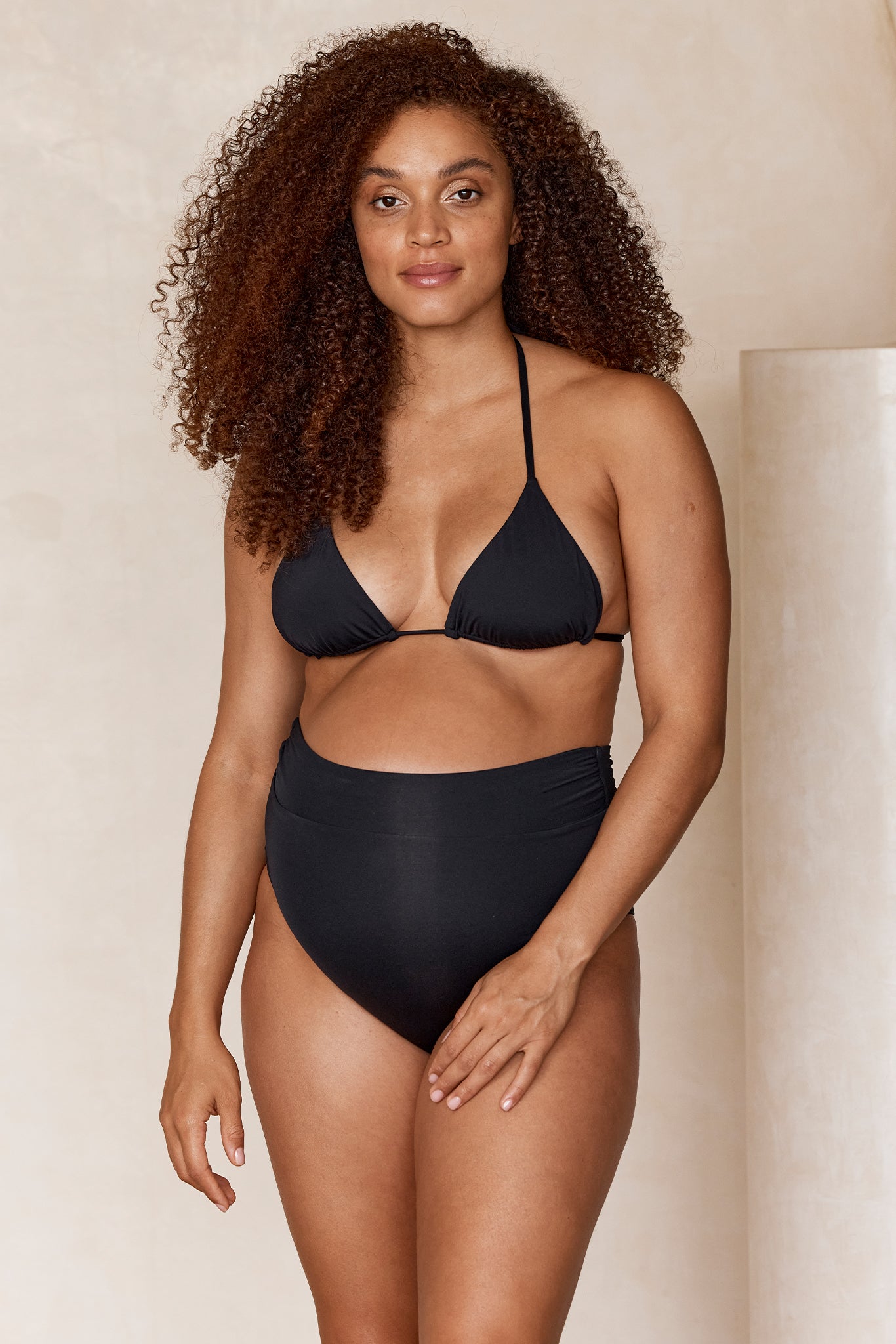 Woman with curly hair modeling a black triangle bikini top and high-waist bikini bottoms, neutral studio backdrop.
