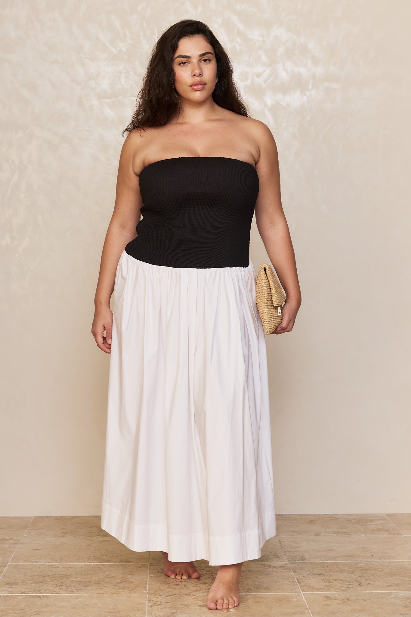 Plus-size model in a black strapless smocked top and full white maxi skirt, holding a woven clutch, barefoot.