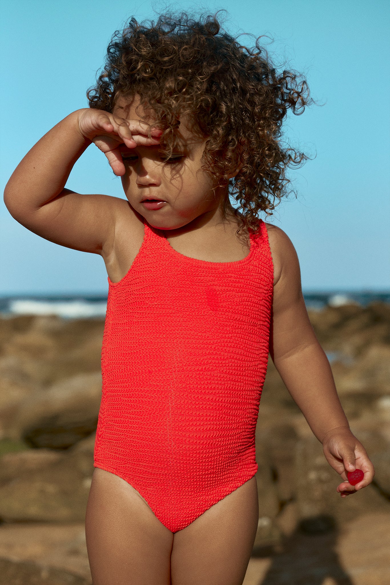 Toddler in textured red swimsuit shielding her eyes from the sun on a rocky beach, holding a small red object.