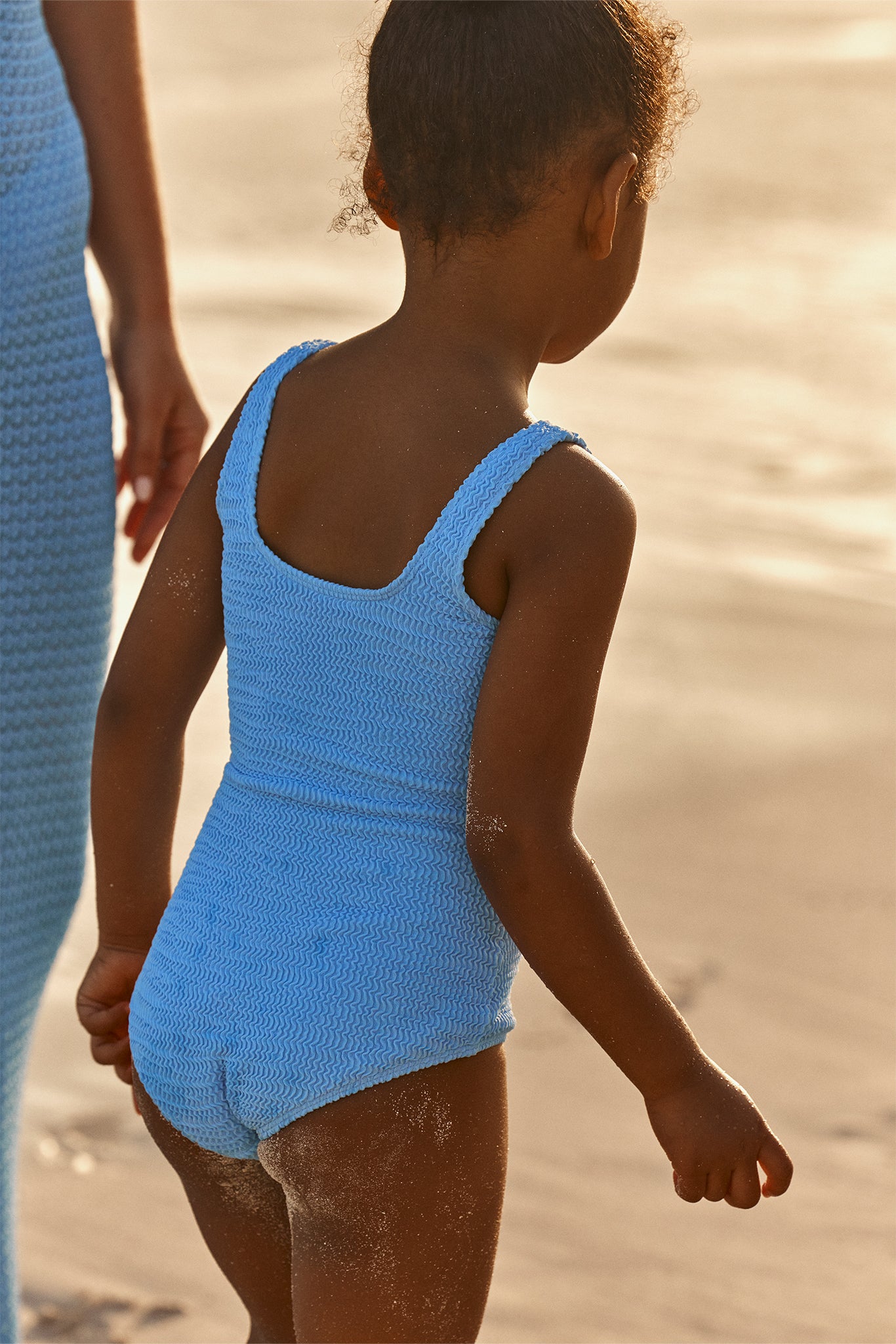 Young child in textured light-blue one-piece swimsuit walking on sandy beach, viewed from behind.