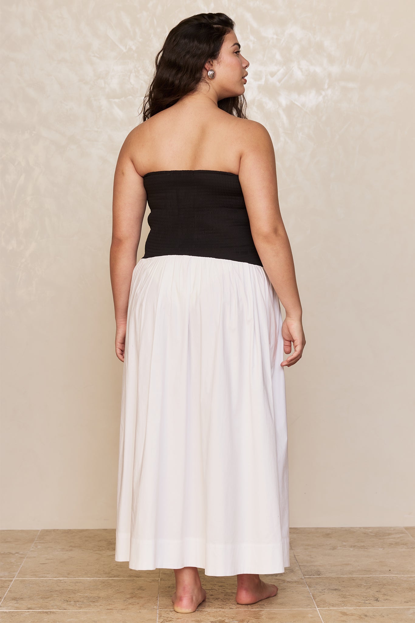 Young woman standing with her back to camera wearing a strapless black top and long white skirt, barefoot on tile floor