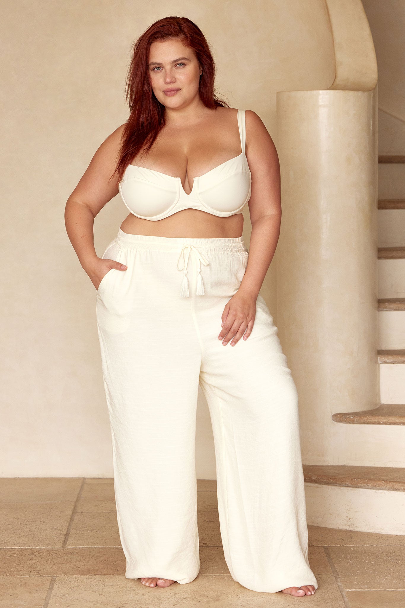 Plus-size woman wearing cream underwire bra and wide-leg drawstring linen pants, standing with one hand in pocket