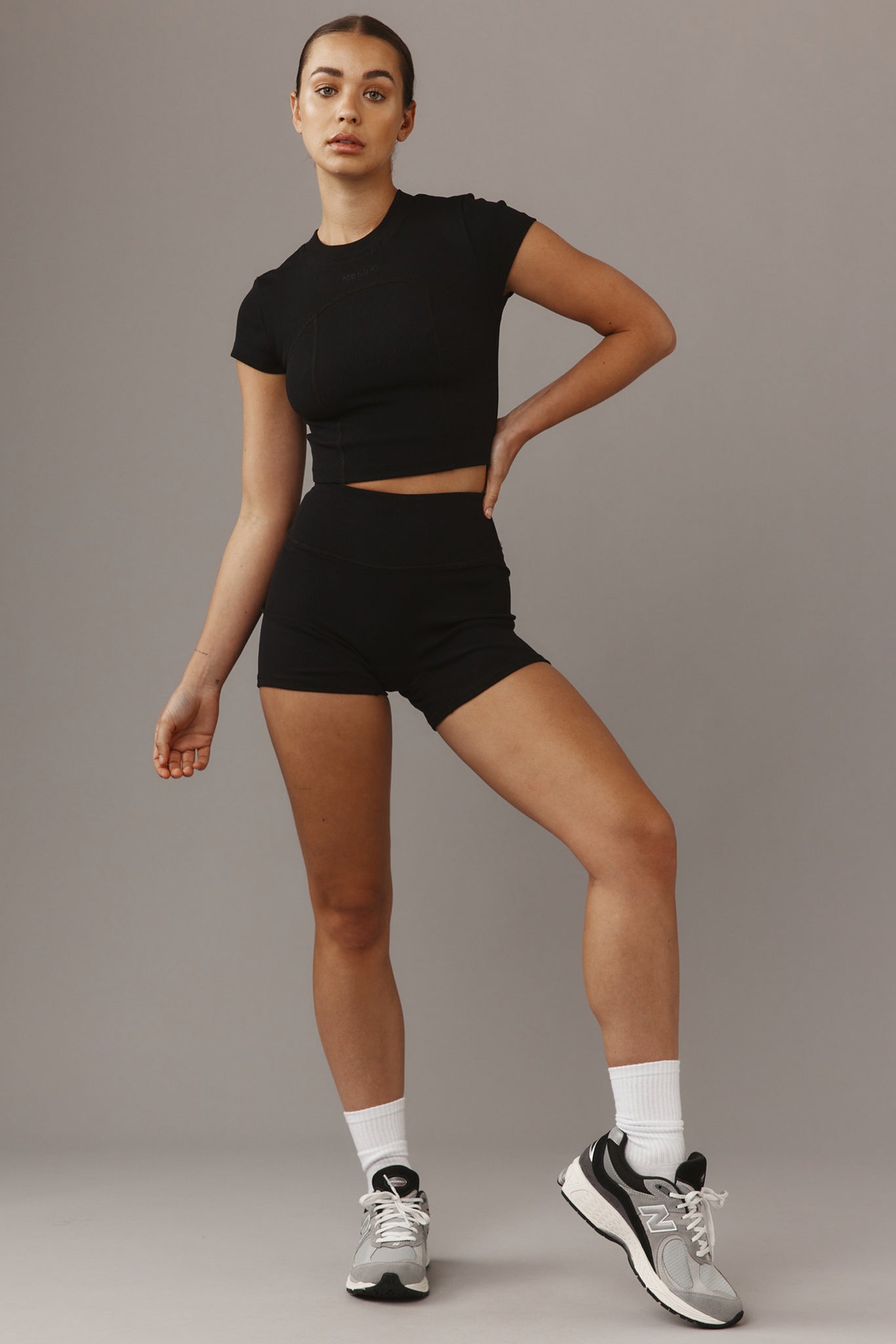Woman wearing black crop top and high-waist shorts with gray sneakers, standing with one hand on her hip against a neutral studio background.