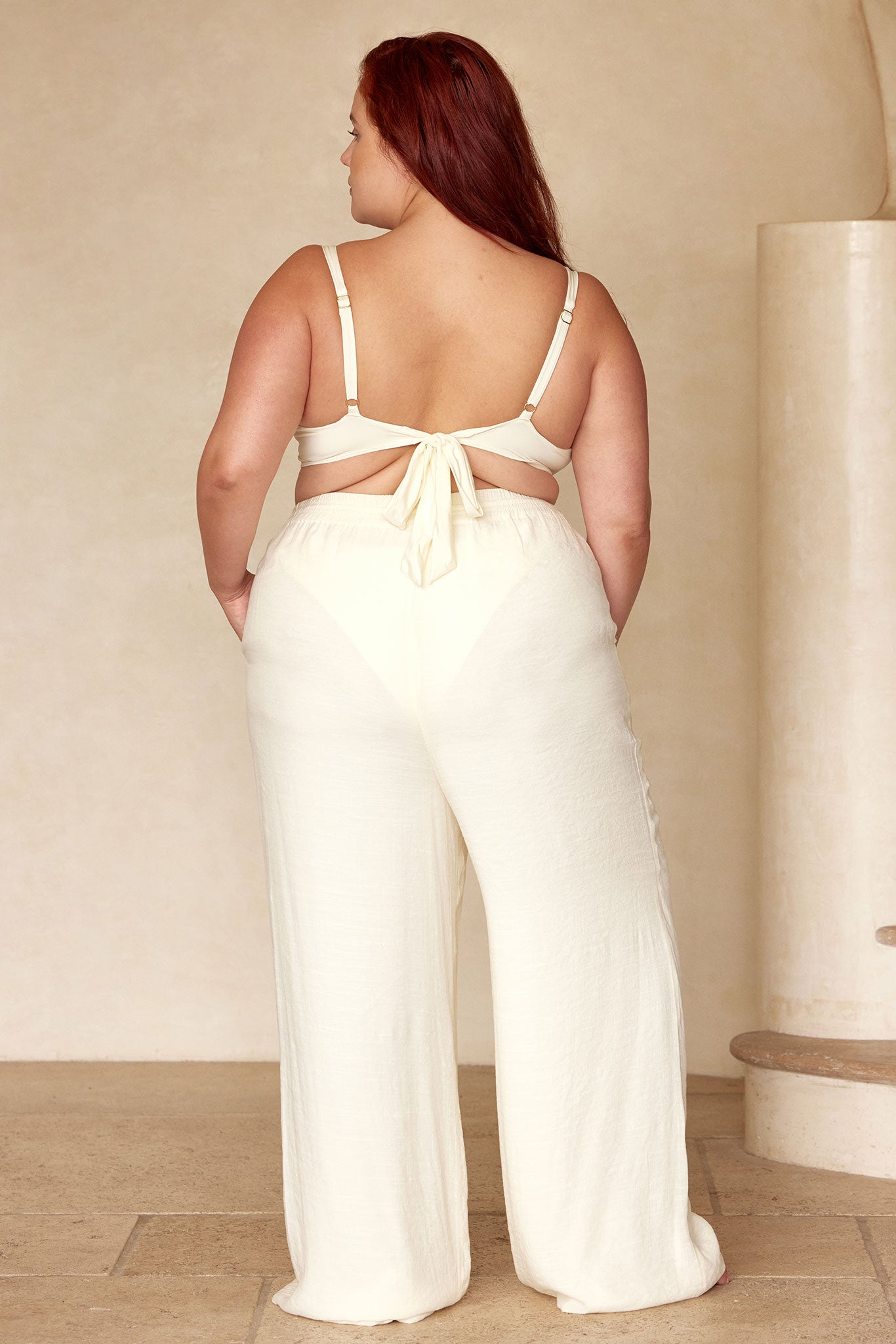 Plus-size woman standing with her back to camera wearing a cream tie-back crop top and high-waist wide-leg trousers in a neutral studio.