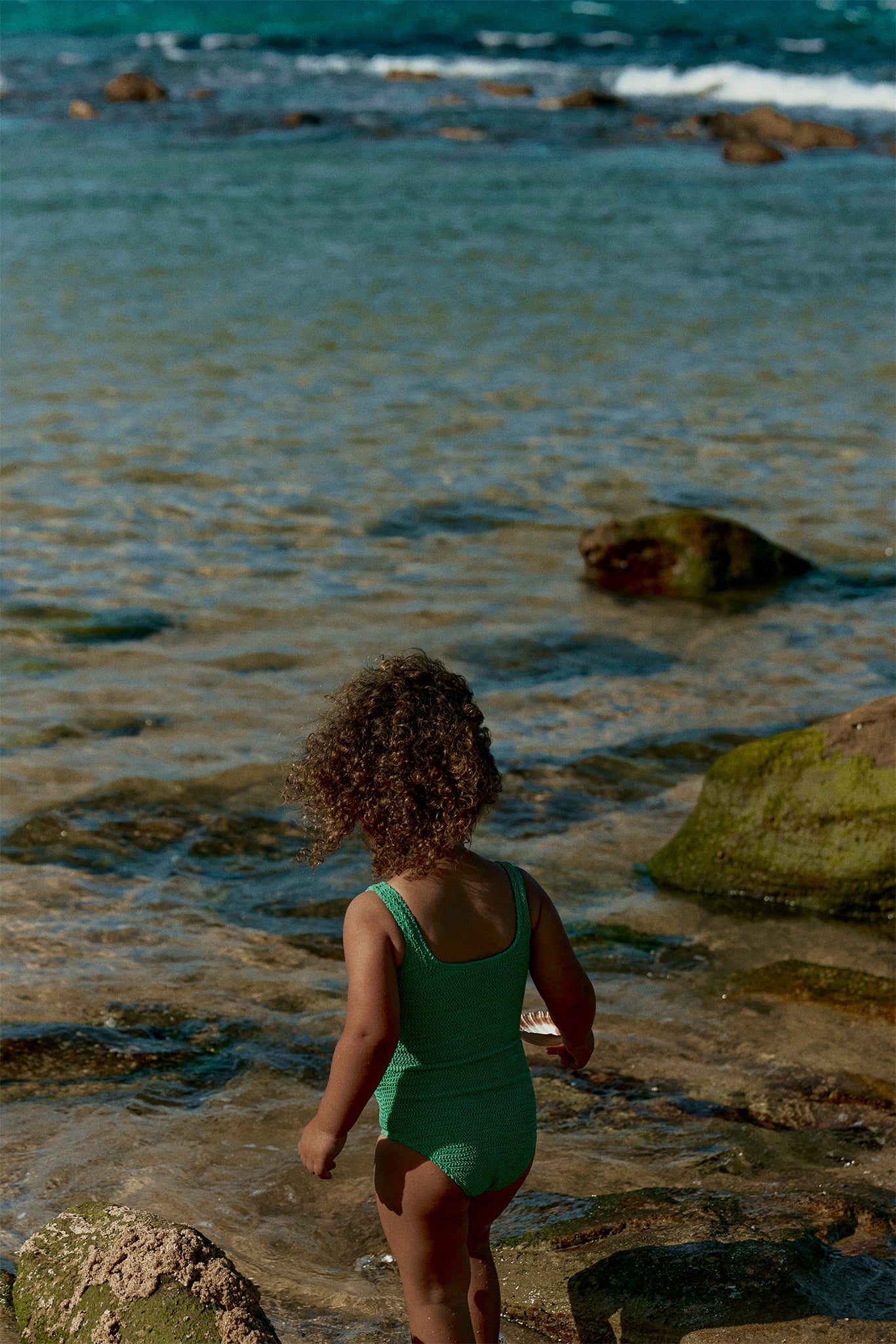 Young child with curly hair in a green swimsuit standing at rocky shoreline, back to camera, holding a white shell